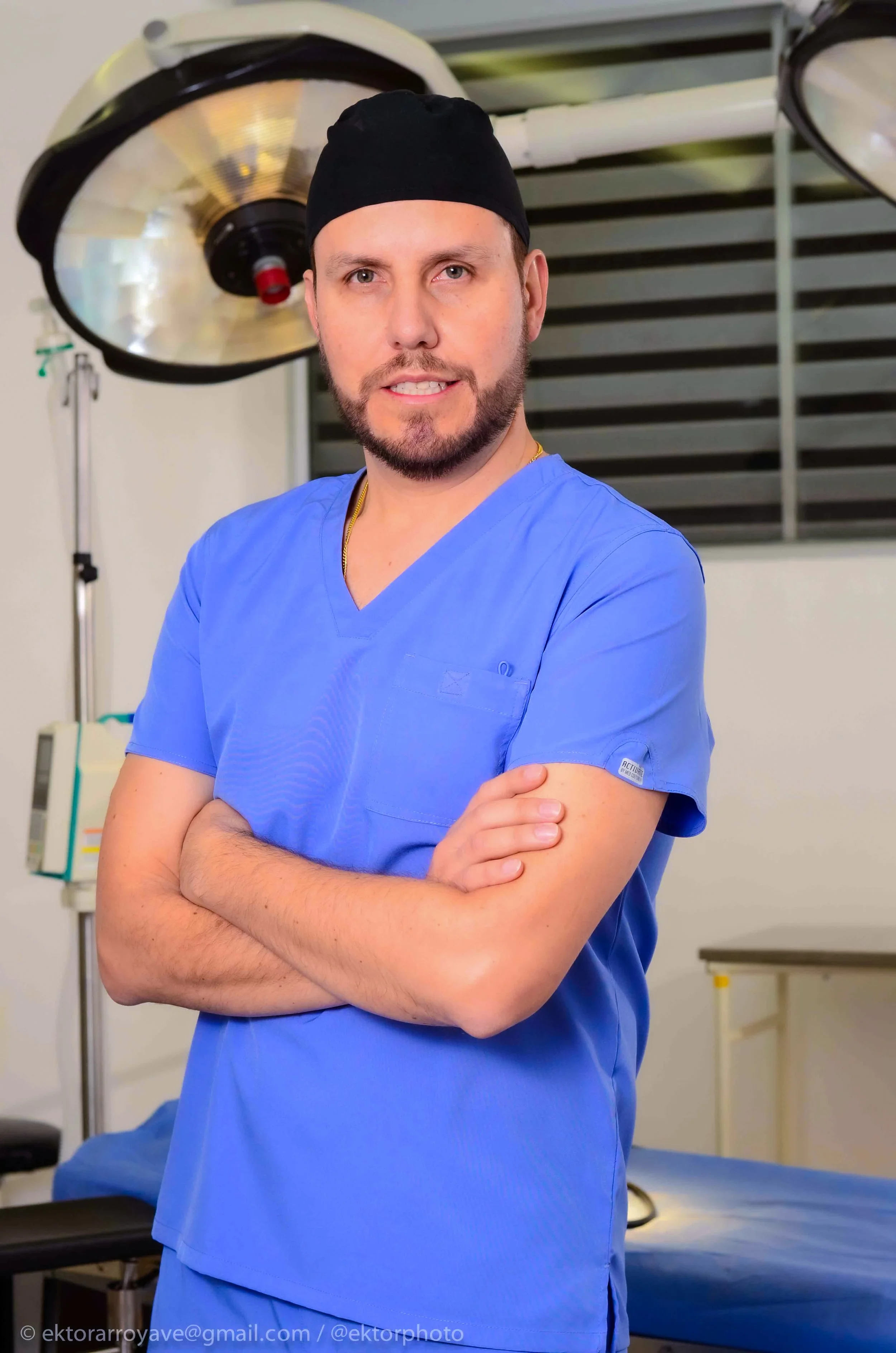 Médico con bata azul, brazos cruzados, en un entorno de quirófano, con equipo médico y luces quirúrgicas de fondo.
