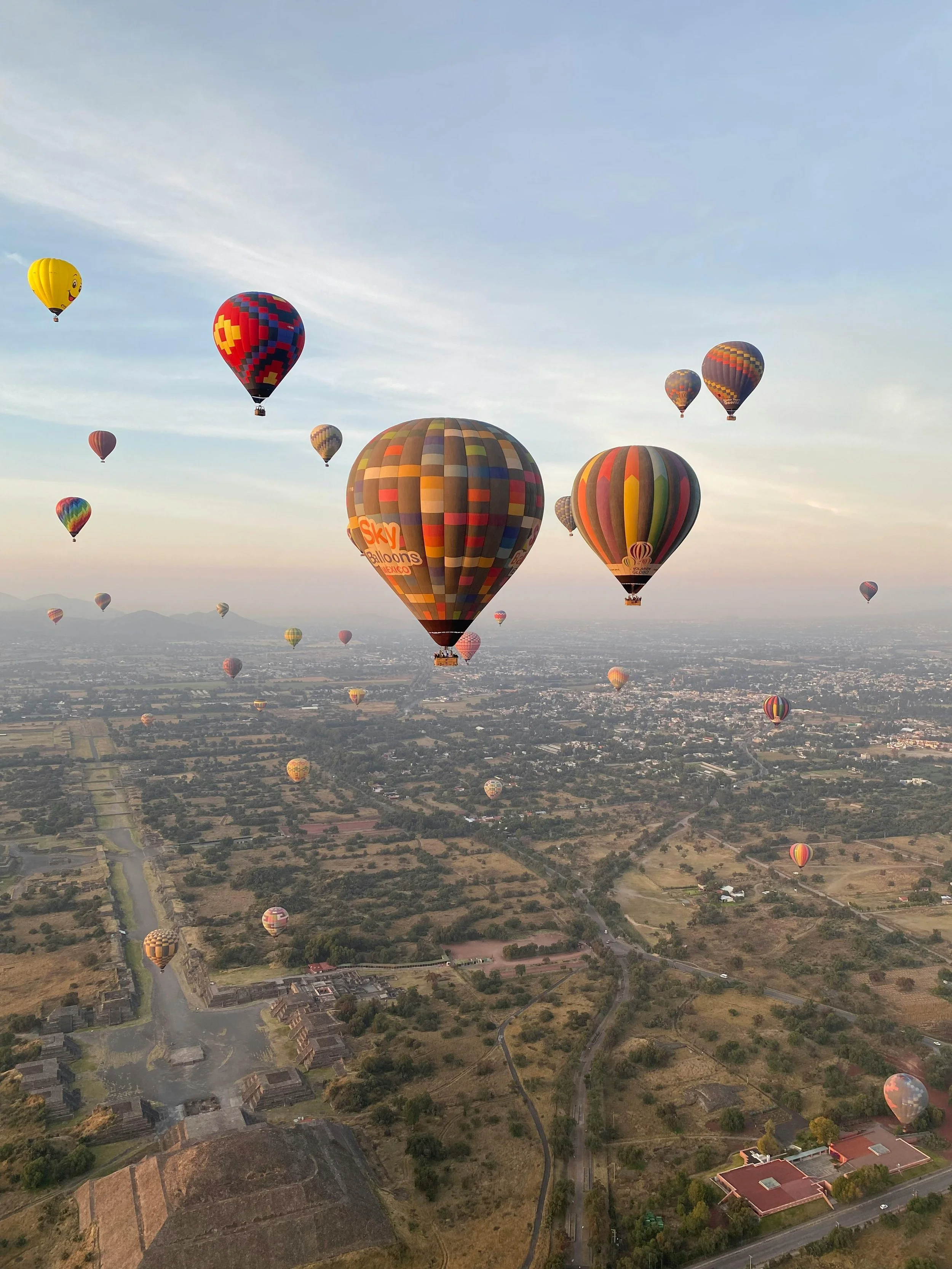 Multiple colorful hot air balloons floating over a landscape with fields, roads, and buildings.