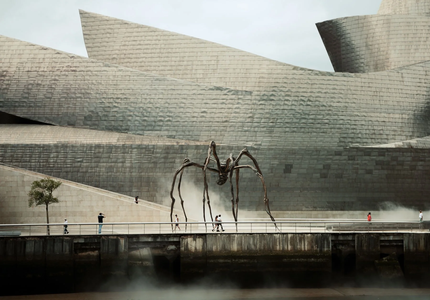 Large spider sculpture in front of a modern, metallic building with people walking on a promenade.