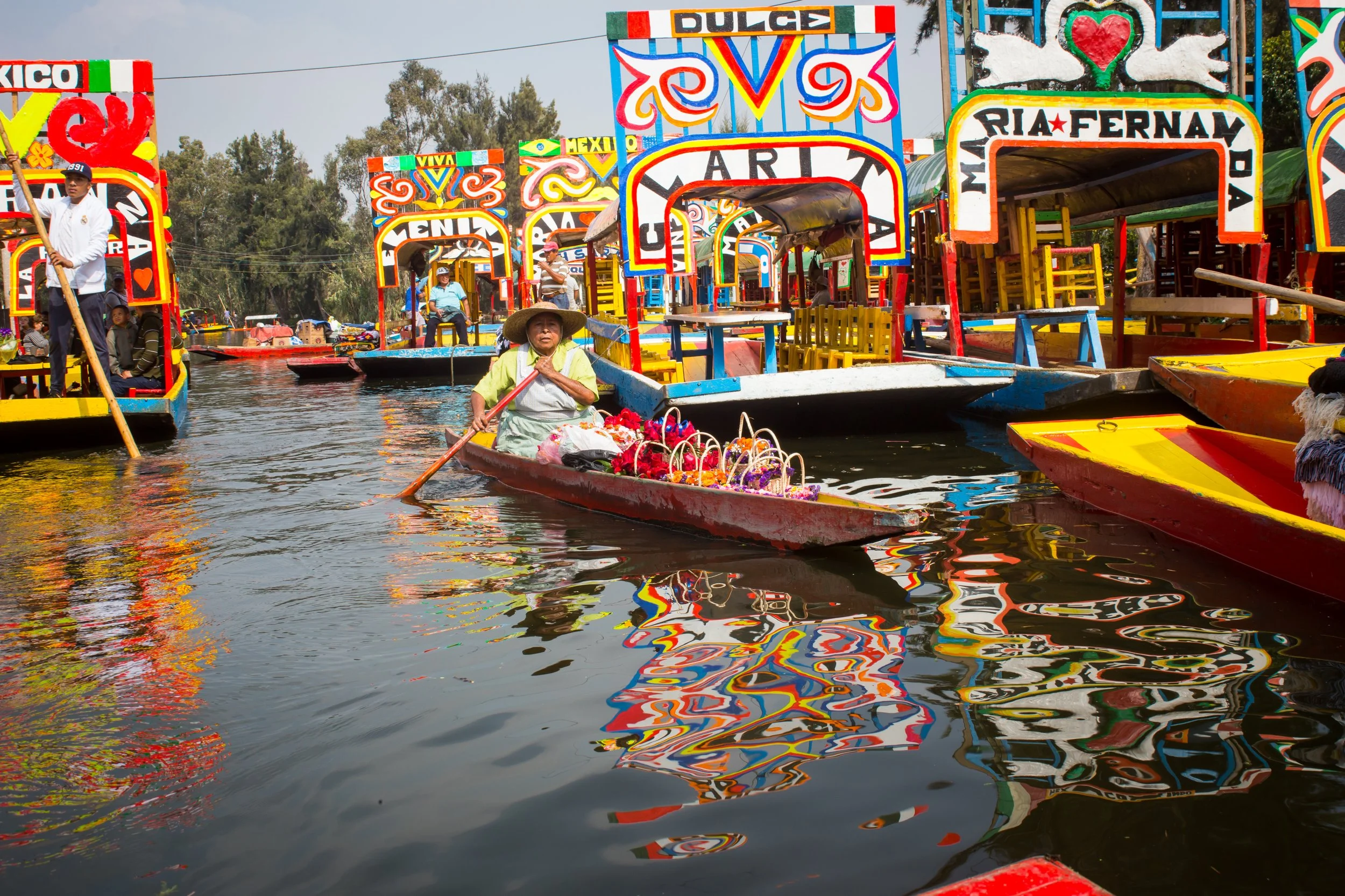 Colorful floating boats on water with people, decorated with vibrant patterns and signs, during a Mexican cultural event or festival.