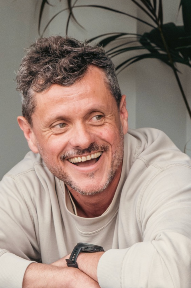 A smiling middle-aged man with gray and brown hair, wearing a light-colored sweatshirt, sitting indoors with a smile, in front of a large plant.