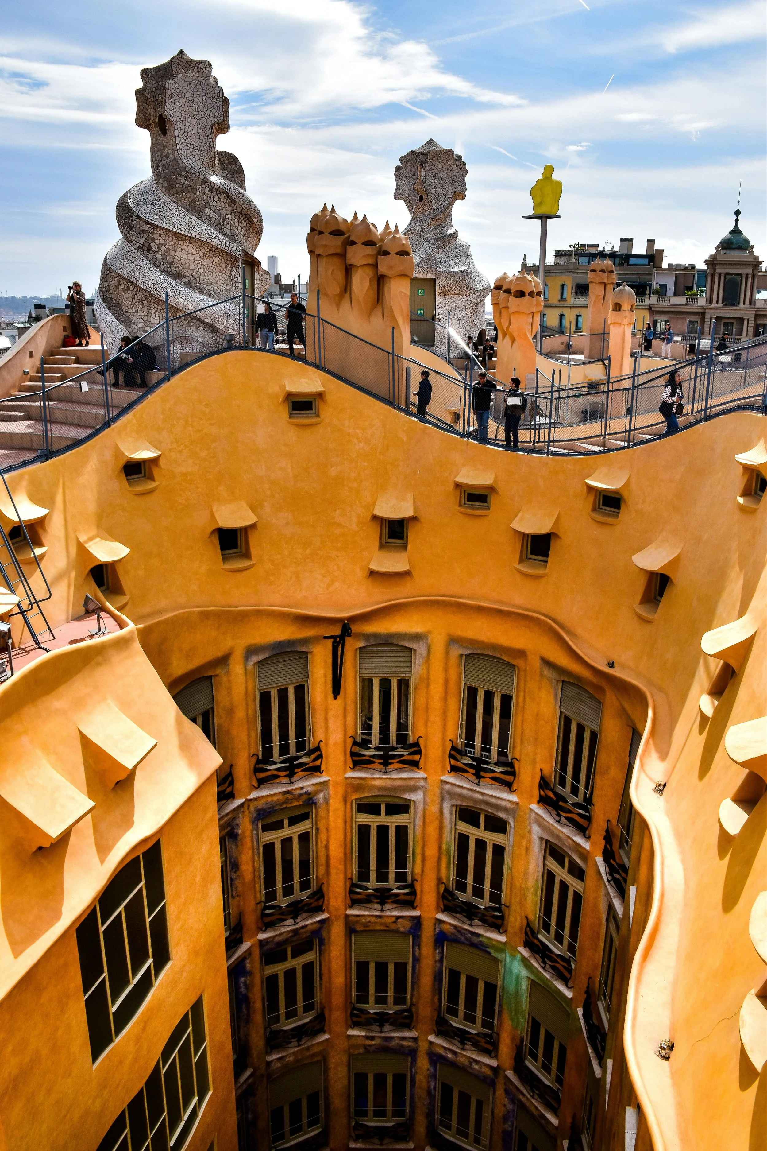 View of the rooftop terrace at Casa Milà in Barcelona, showing its distinctive undulating architecture, decorative chimneys, and sculptures, with people walking around and the city skyline in the background.