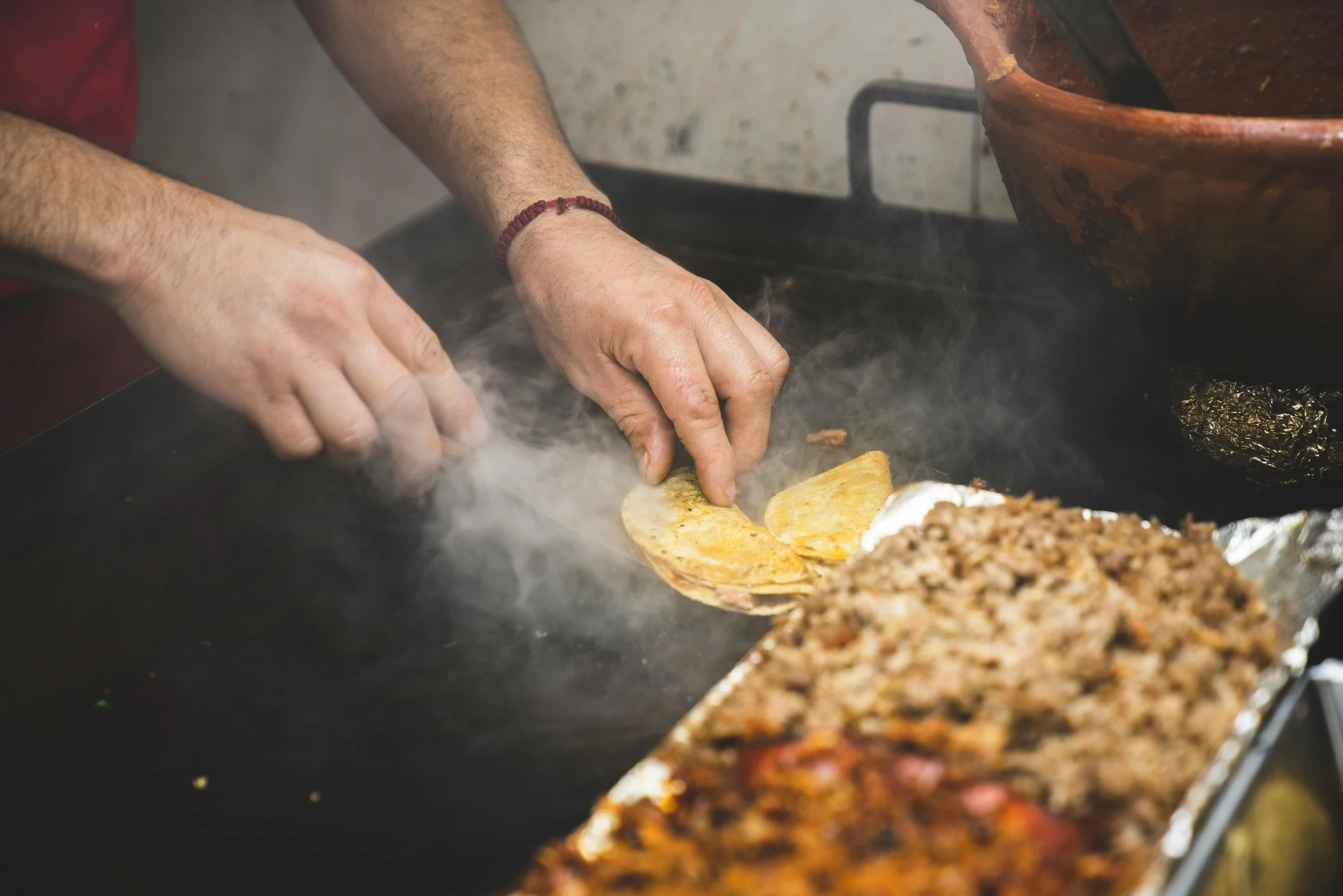 Hands preparing tacos on a stovetop with sizzling ingredients, including ground meat in the foreground.