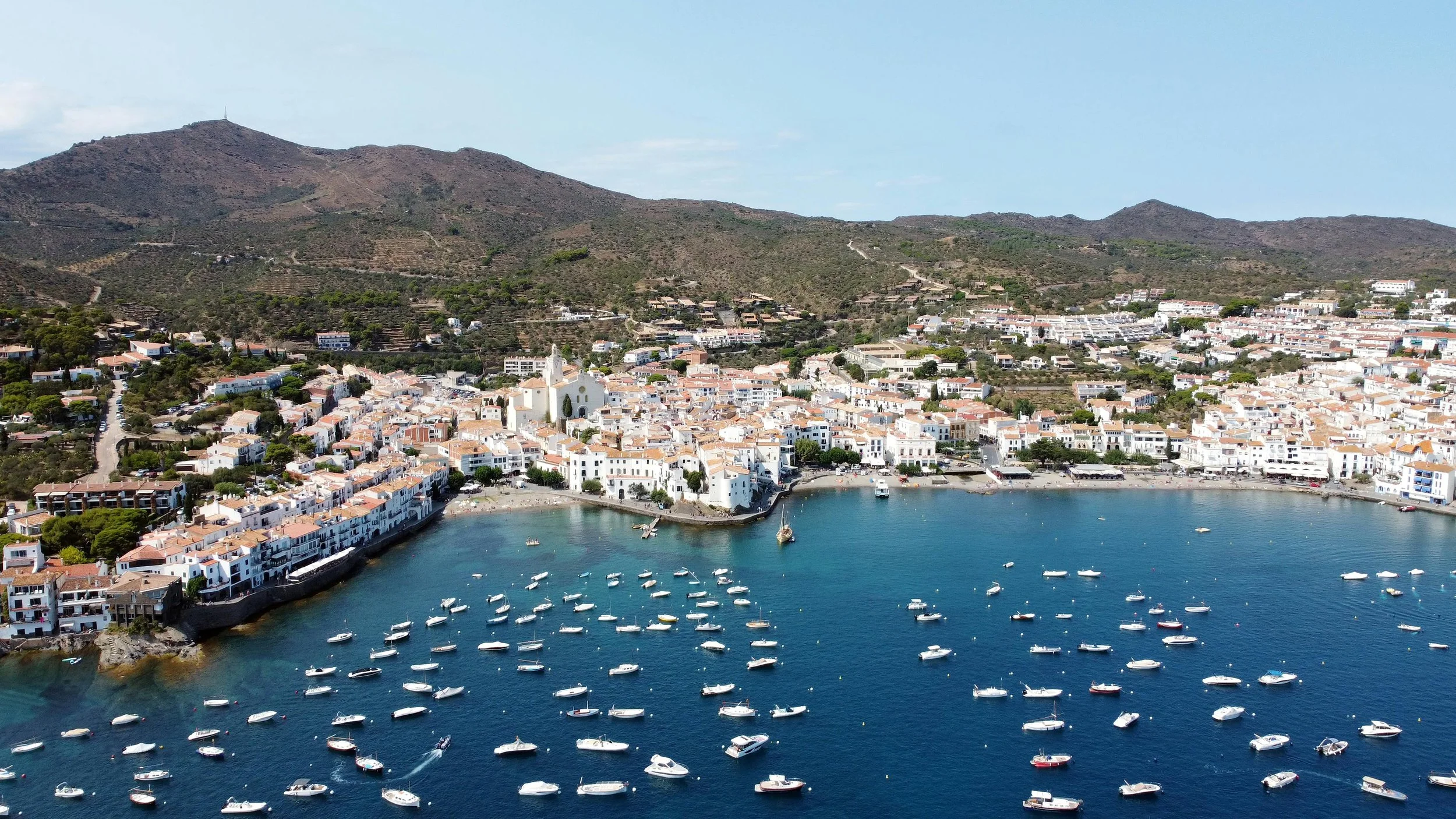 Aerial view of a coastal town with white buildings and a marina filled with boats, surrounded by mountains.
