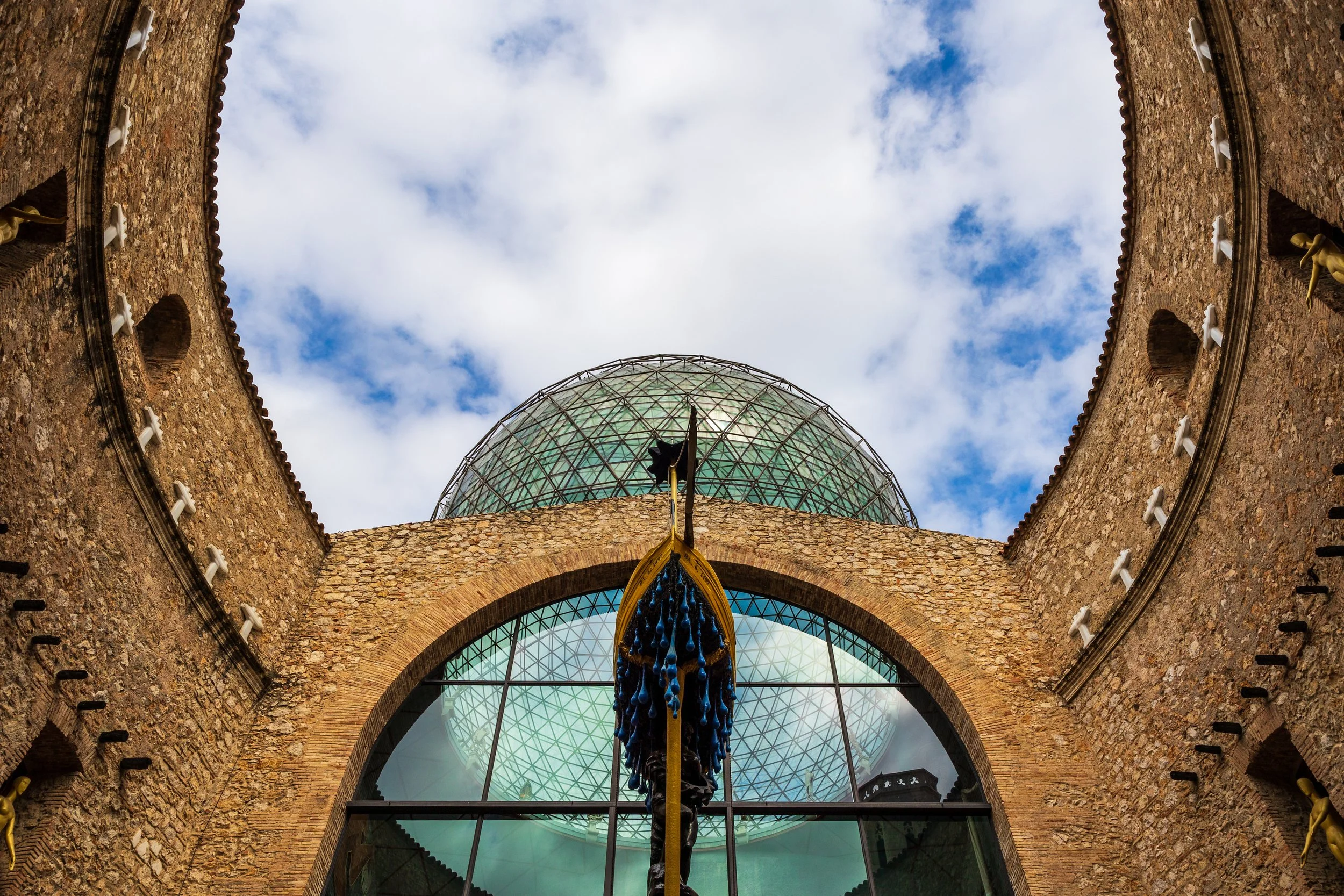 Looking up at a historic brick building with a glass dome and a statue of a figure holding a flag in front of the large window, with a partly cloudy sky above.