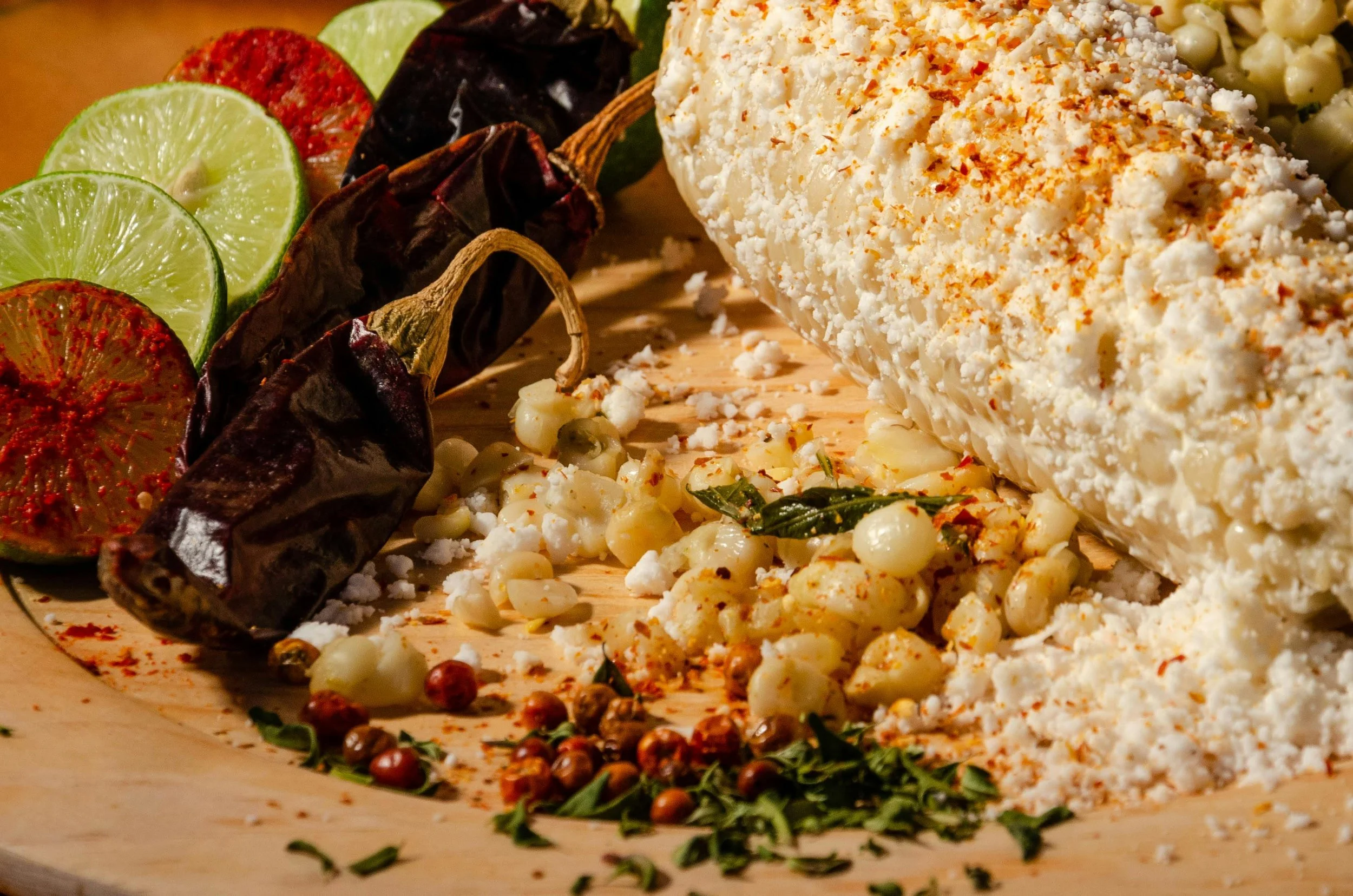 Close-up of fresh ingredients on a wooden cutting board, including limes, roasted chili peppers, crushed white cheese, and small white pearl onions, with herbs and spices sprinkled around.