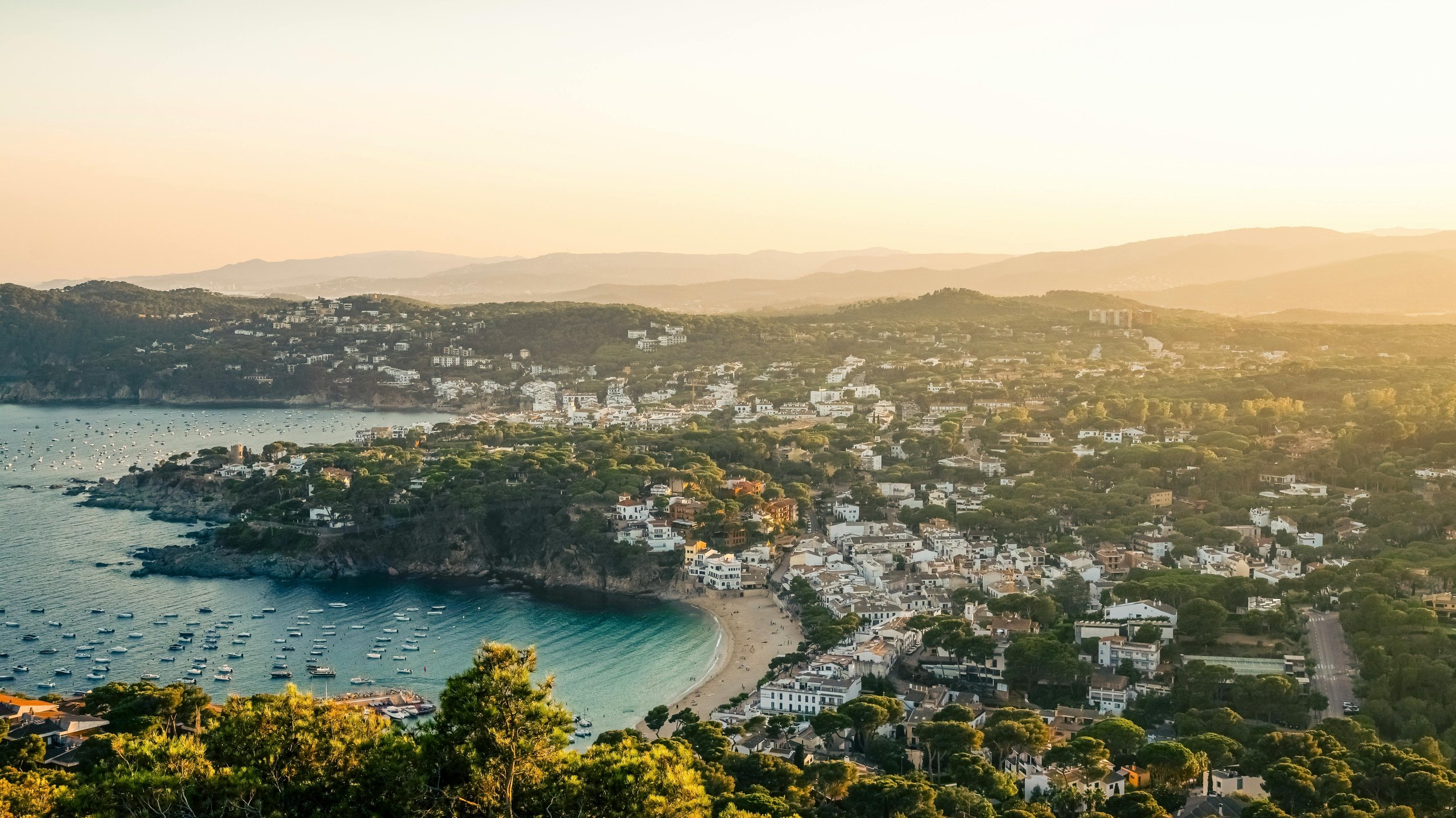 Aerial view of a coastal town with a crescent-shaped beach, boats in the bay, and houses on hills during sunset.