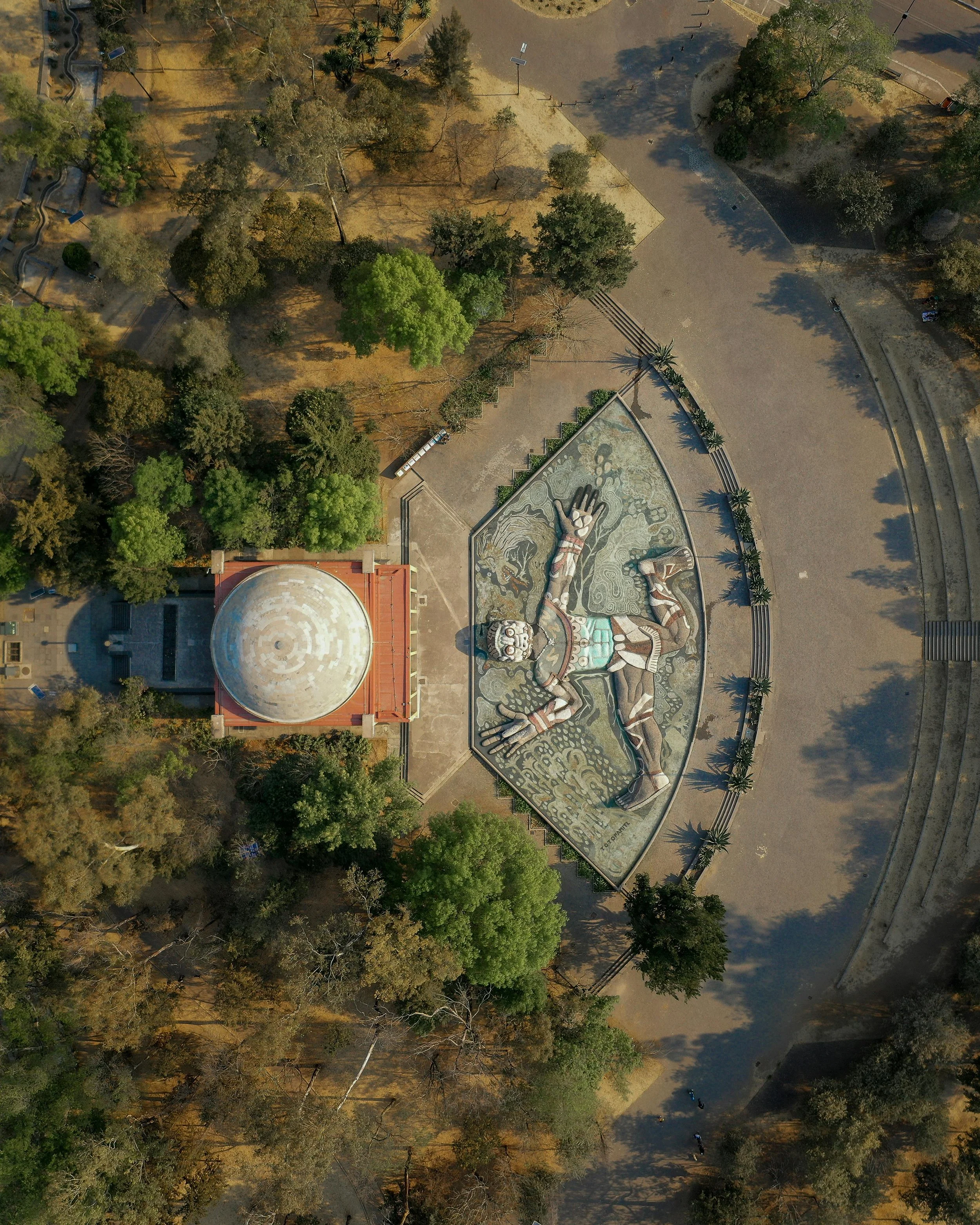 An aerial view of a park featuring a large mosaic mural of a reclining person with outstretched arms, surrounded by trees and pathways.