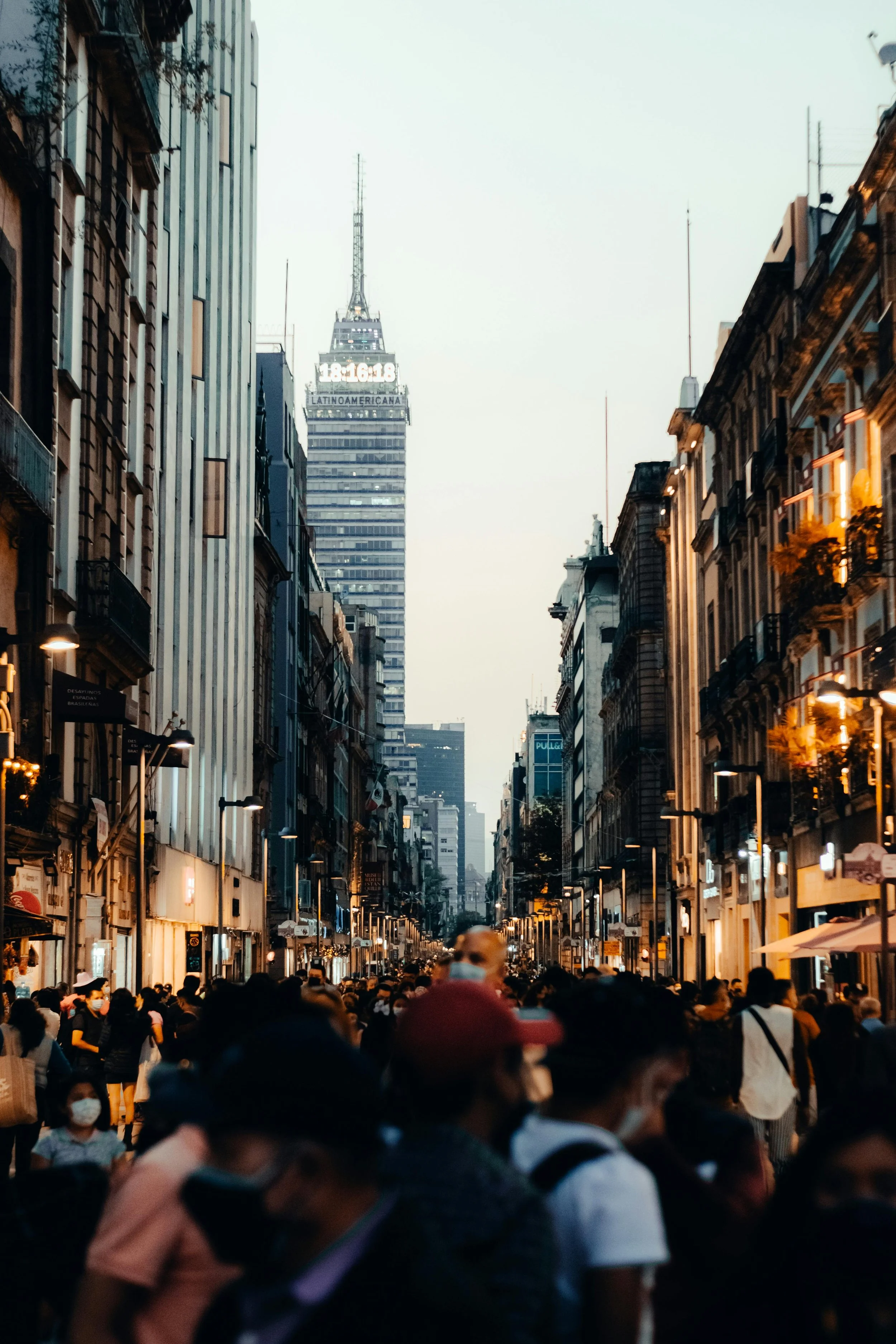 A crowded city street during dusk with tall buildings on either side and a prominent skyscraper in the background. People are wearing masks and walking along the street, which is illuminated by street lights.