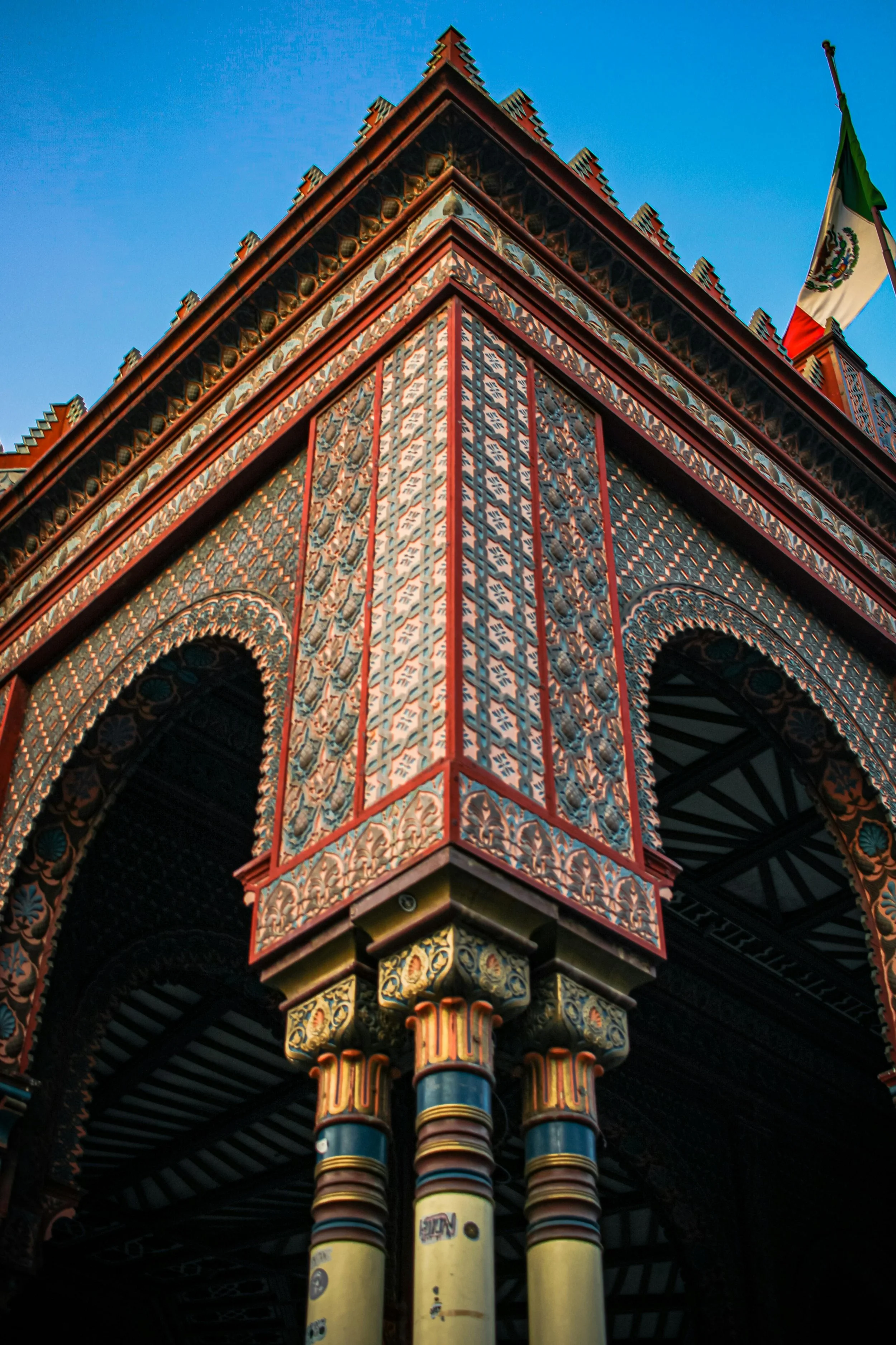 Close-up of an ornately decorated structure with patterned walls and columns, topped with a flag against a clear blue sky.