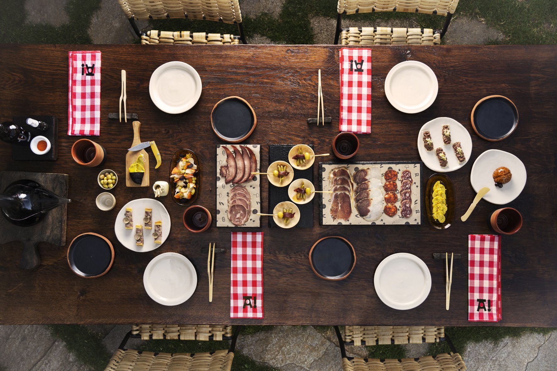 Overhead view of a wooden dining table set for a meal with assorted appetizers, small plates, bowls of sauces, wine glasses, chopsticks, and red checkered napkins, surrounded by wicker chairs.