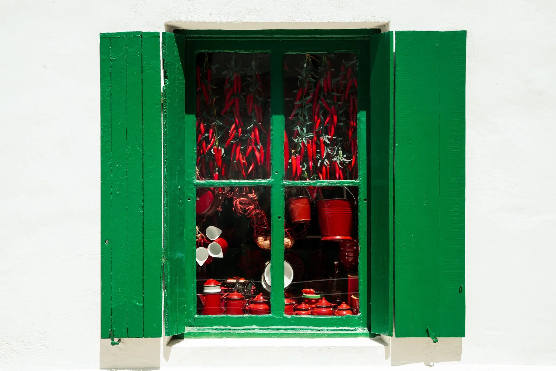 Green window with shutters displaying red chili peppers and red ceramic kitchenware inside.