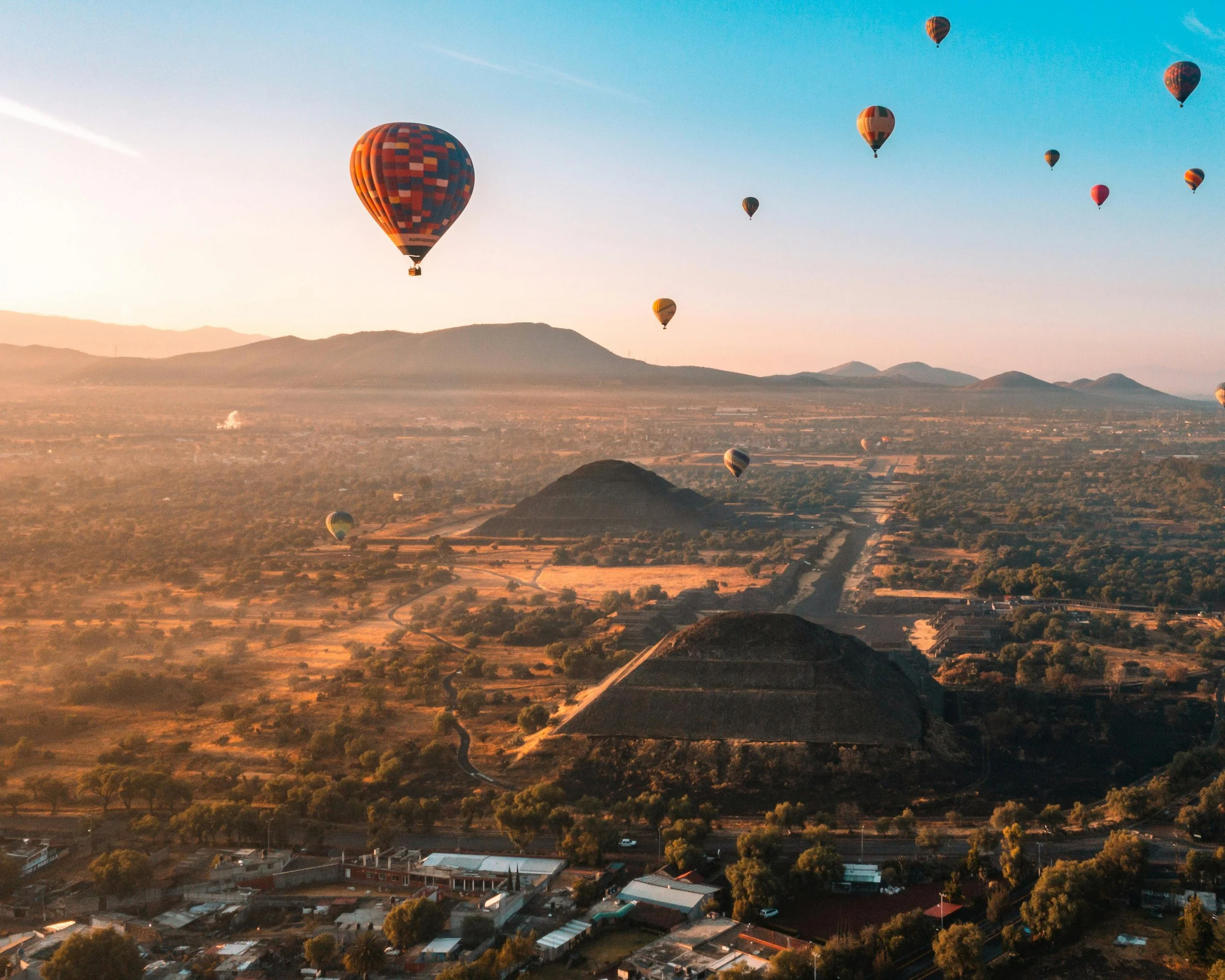 Multiple hot air balloons floating over an expansive landscape with ancient pyramids and low mountains at sunrise.