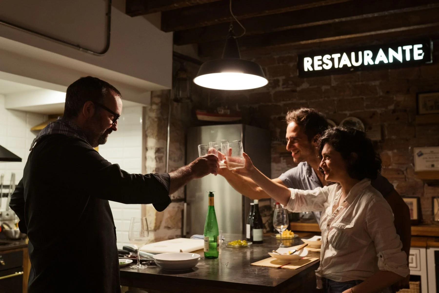 Three people toast glasses in a restaurant kitchen, smiling, with a sign that says 'RESTAURANTE' on a brick wall.