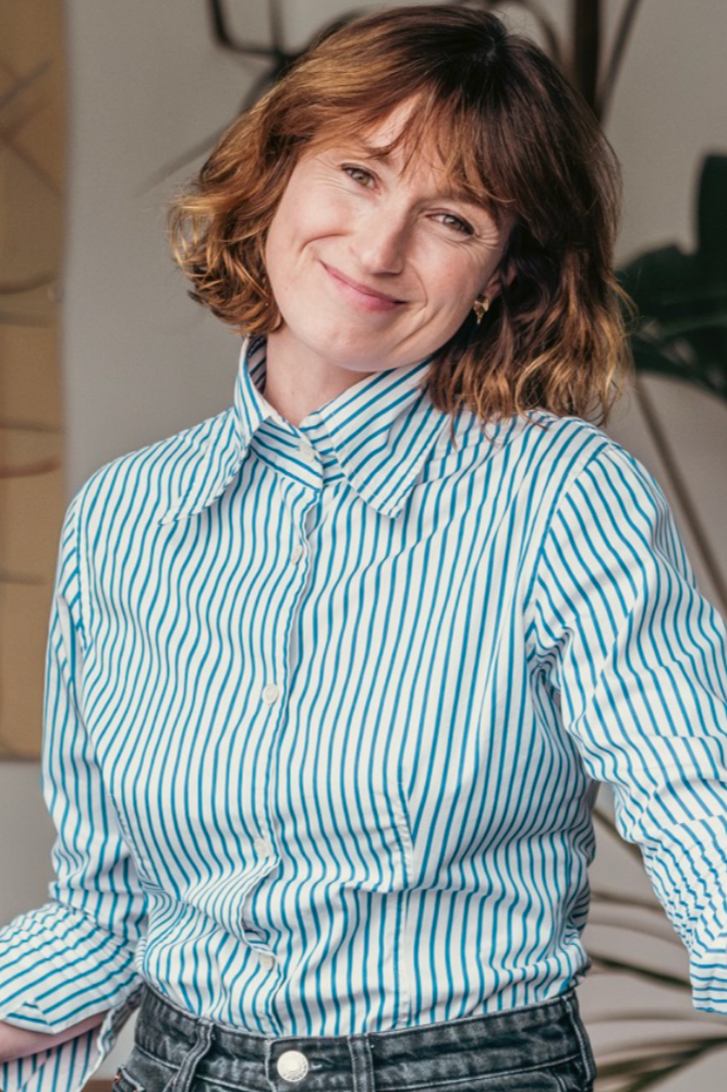 A woman with short wavy brown hair is smiling and winking at the camera. She is wearing a blue and white striped button-up shirt and black jeans, standing in an indoor setting with plants and shelves in the background.