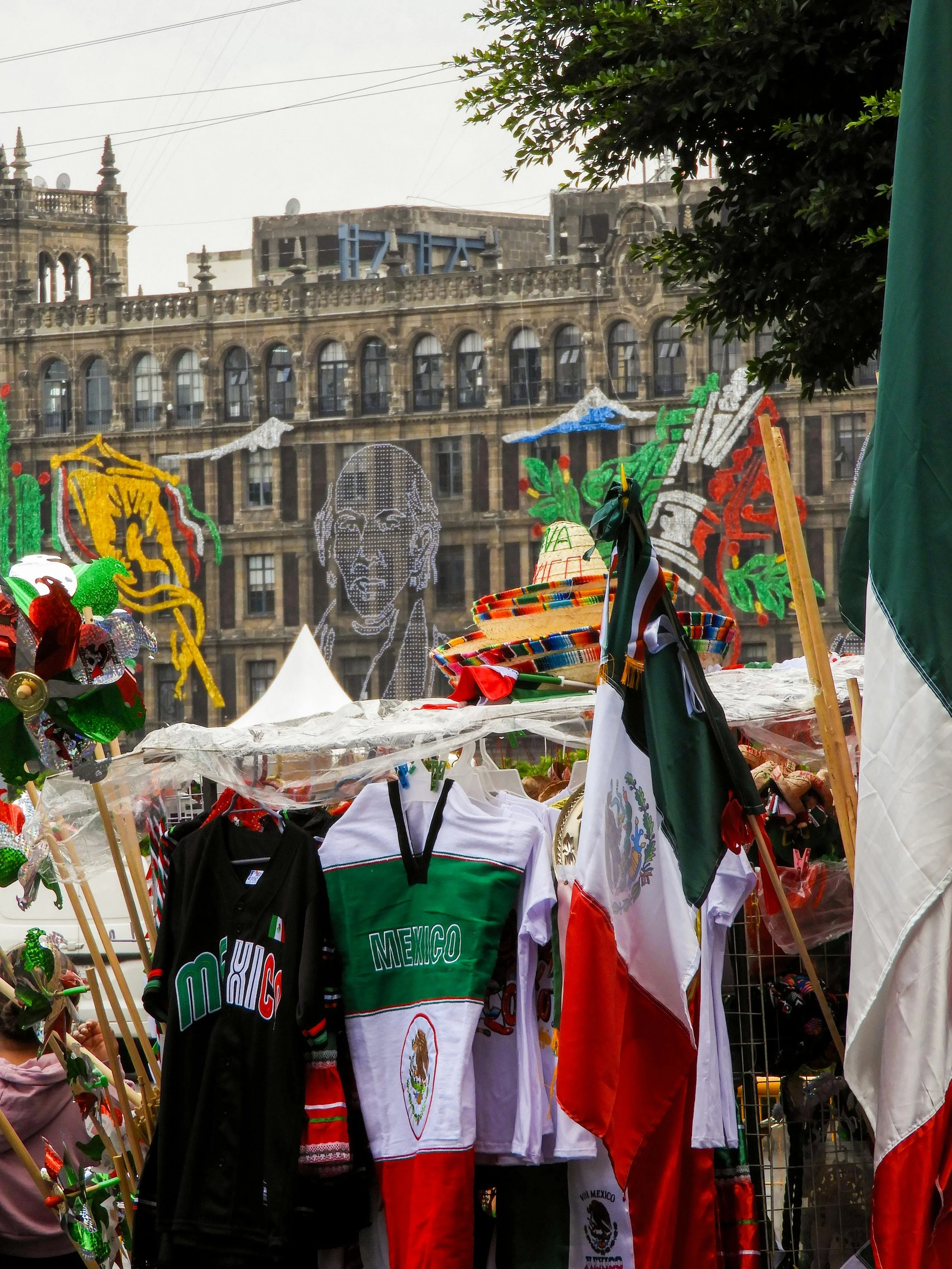 Market stall with Mexican and other flags, hats, and souvenirs in a city square, with historic building and colorful decorations in the background.