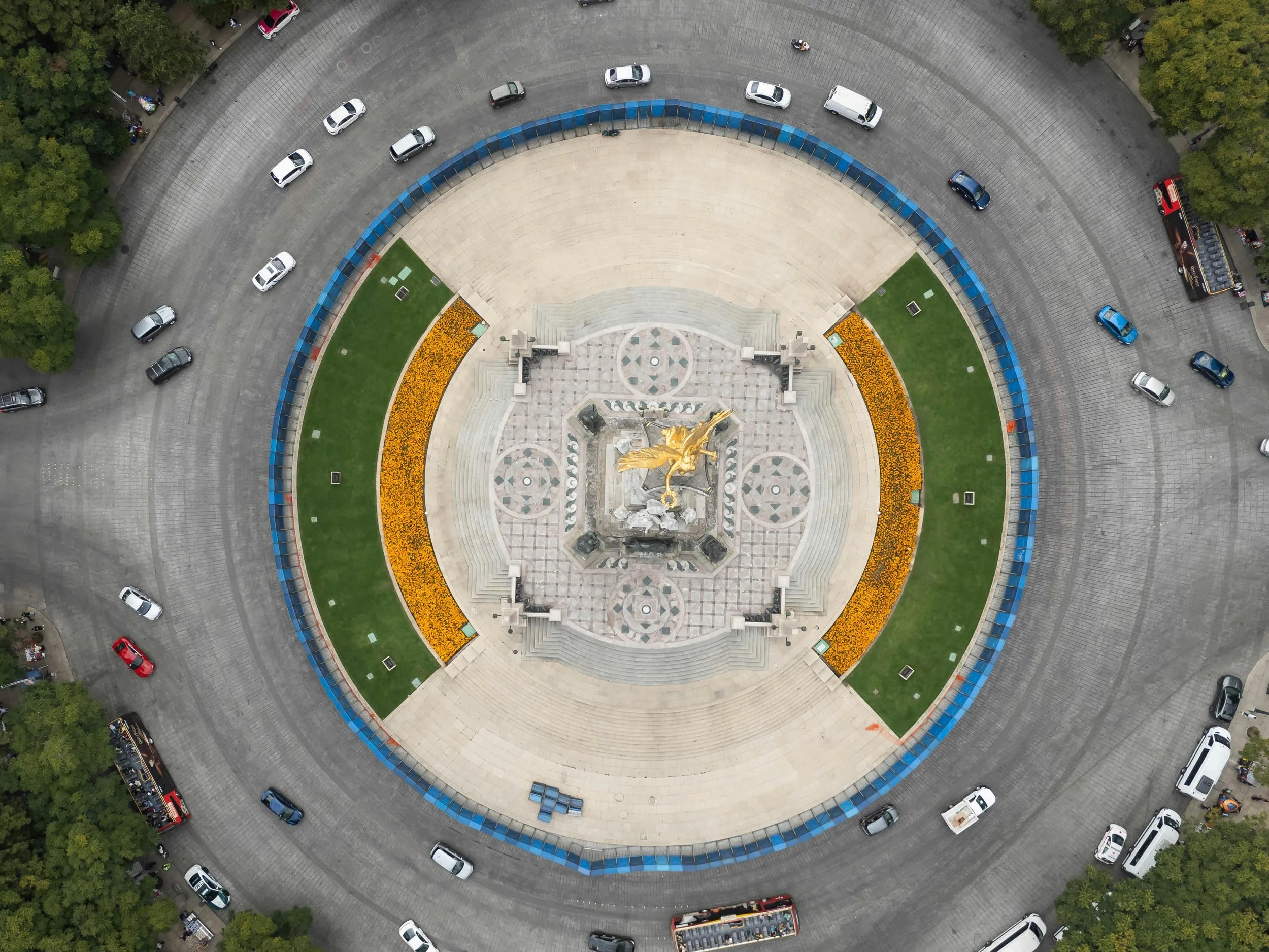 An aerial view of a circular plaza with a gold sculpture in the center, surrounded by landscaped green areas with yellow flowers, paved walkways, and parked cars around the perimeter.