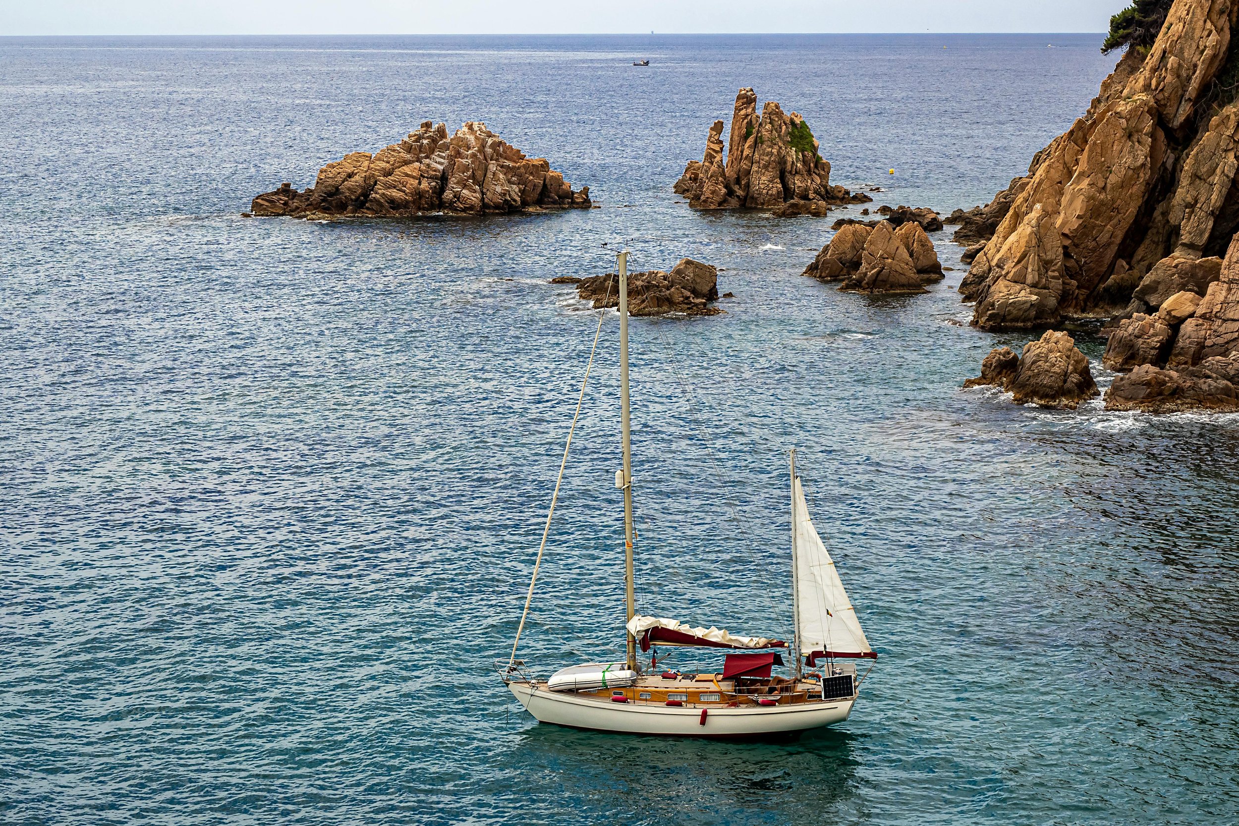 A sailboat with a white sail and solar panel floating near rocky coastline and small rocky islands in the ocean.