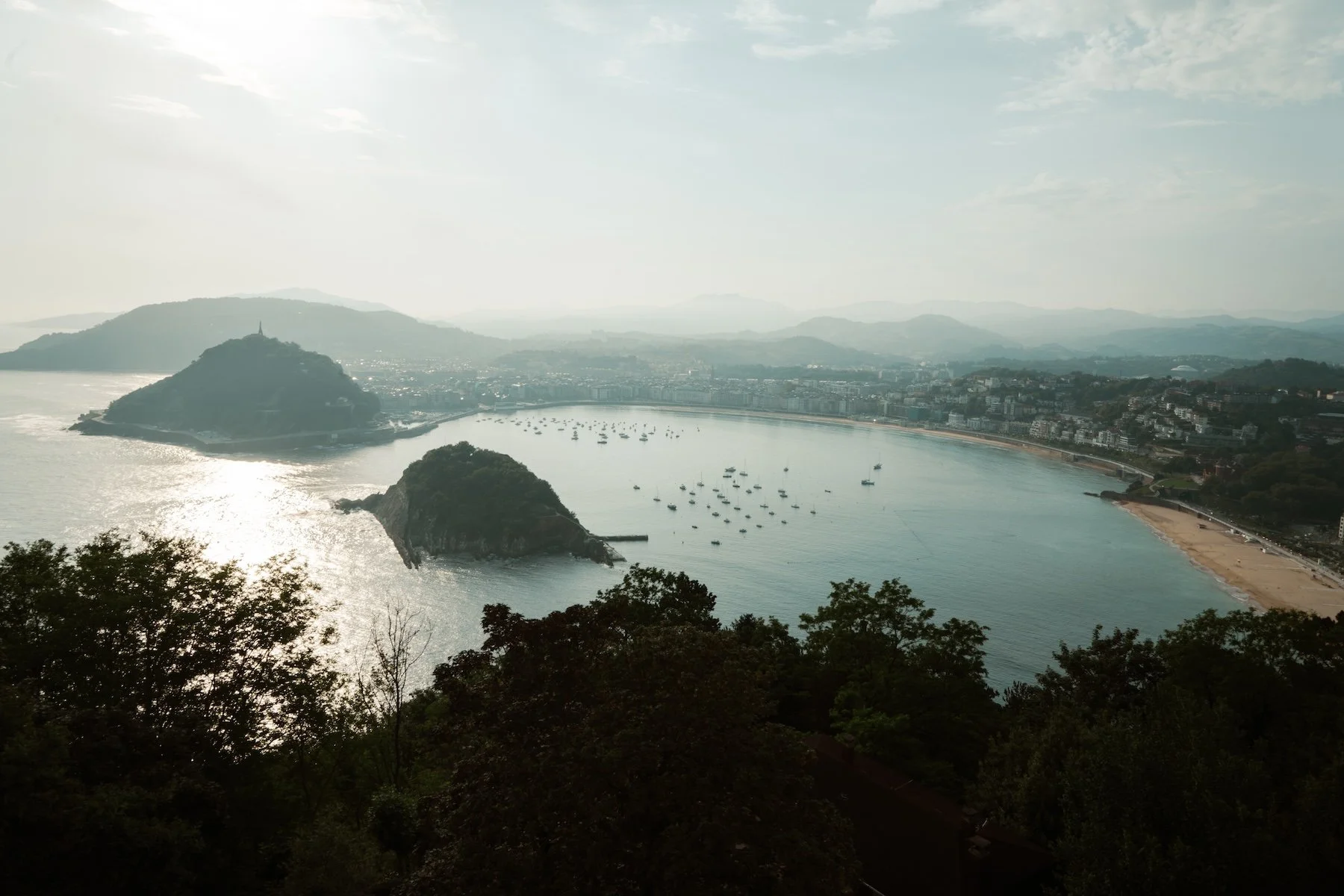A scenic view of a bay with two small islands, boats anchored in the water, a beach along the shoreline, and a city in the distance surrounded by hills and greenery under a cloudy sky.