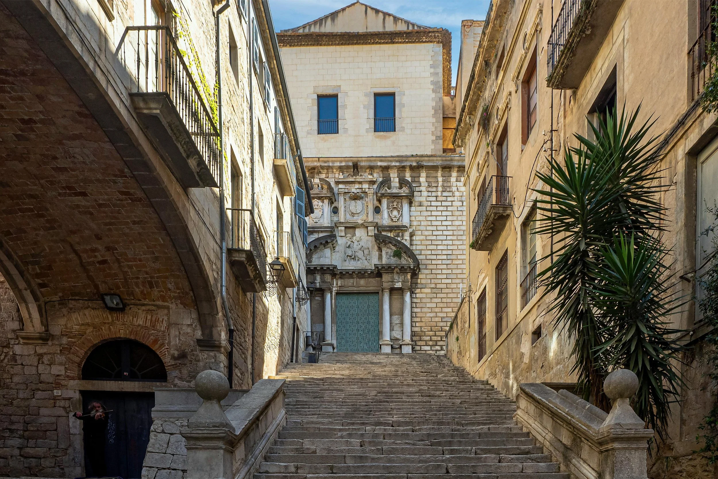 Stone staircase leading up to an ornate historical building with decorative carvings on a facade, flanked by old buildings with balconies, on a sunny day with a blue sky.