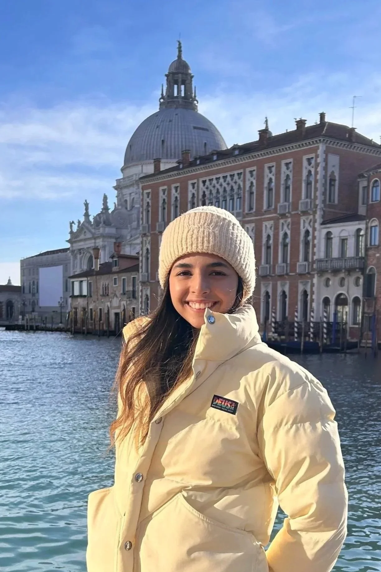 A smiling woman in a yellow jacket and cream knit hat standing by a canal, with historic buildings and a large dome building in the background on a clear day.