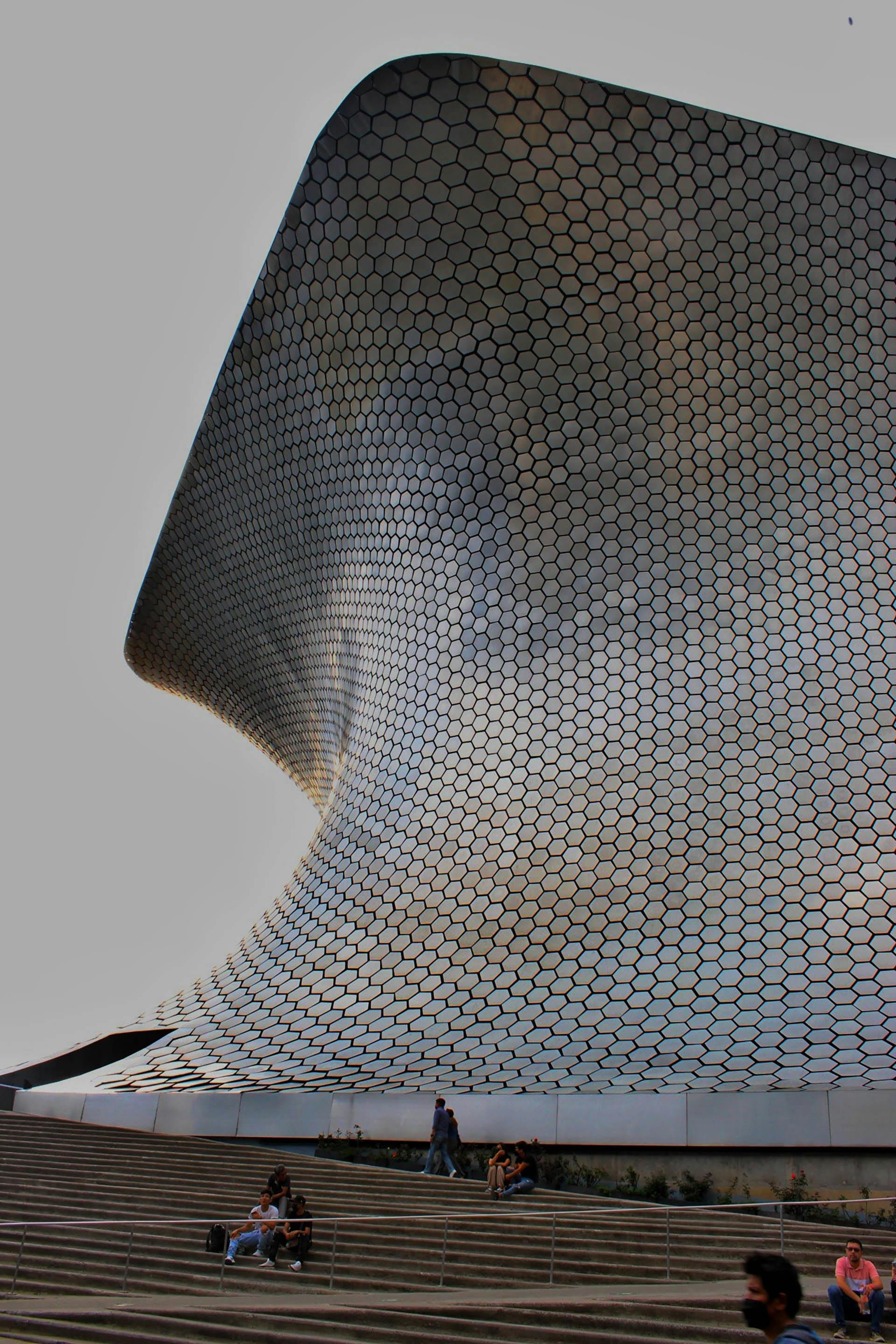 Modern building with curved, honeycomb-patterned exterior and stairs with people sitting and walking outside.