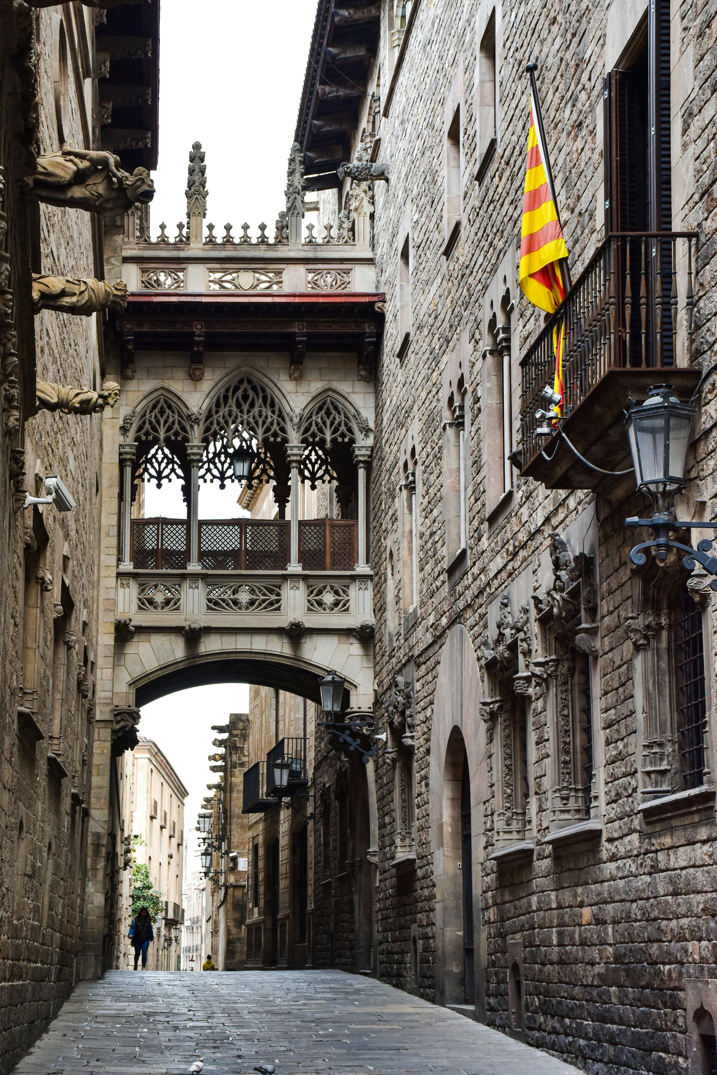 A narrow cobblestone street between old stone buildings with Gothic-style balconies and archways, a Catalan flag hanging from one balcony, and street lamps along the walls.