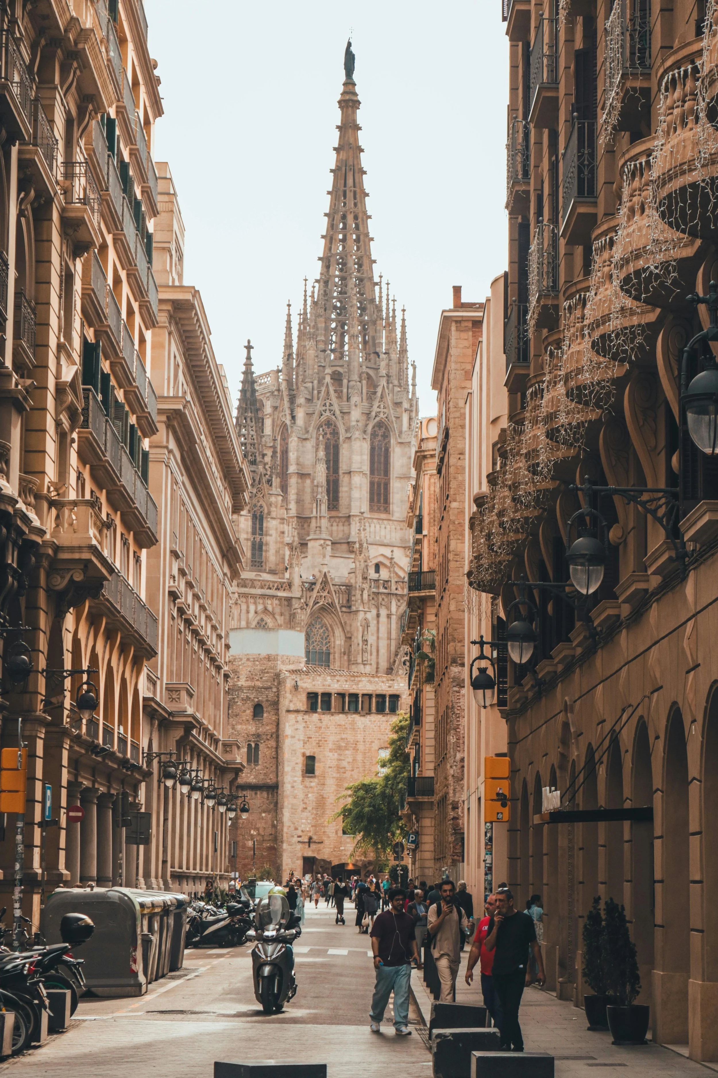 View of a narrow city street with people walking and parked motorcycles, leading to a historic Gothic cathedral in the background with detailed spires and stained glass windows.