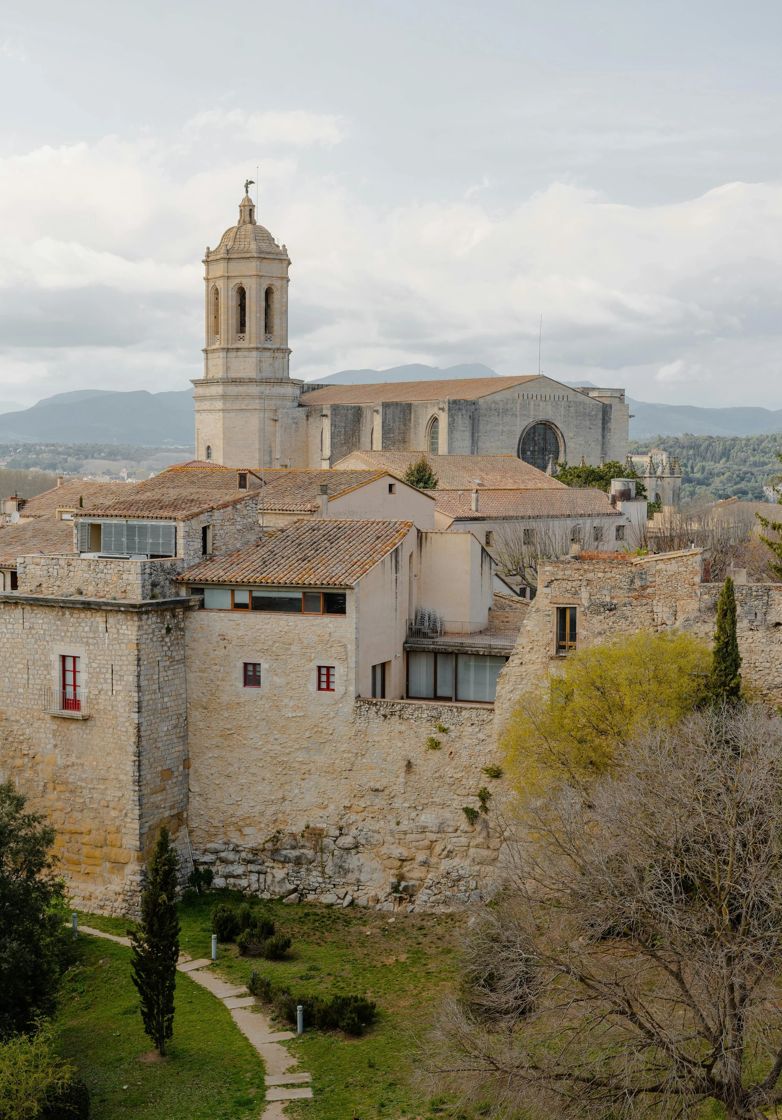 View of a historic European town with a large stone church featuring a tall bell tower, surrounded by old stone buildings, trees, and a lush green park in the foreground.