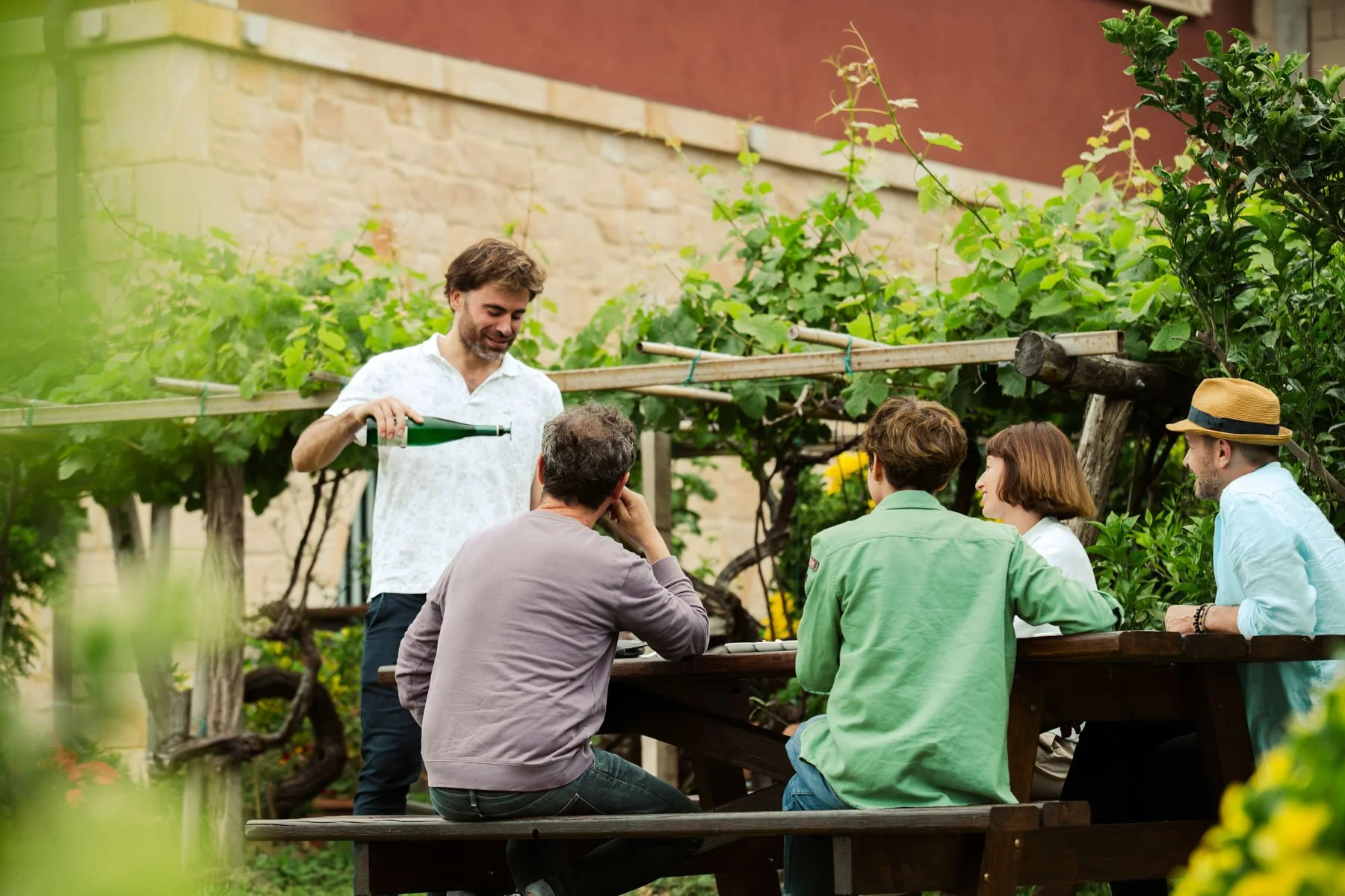 A group of five people sitting around a wooden table in a garden, with one man pouring a drink, and all engaged in conversation.