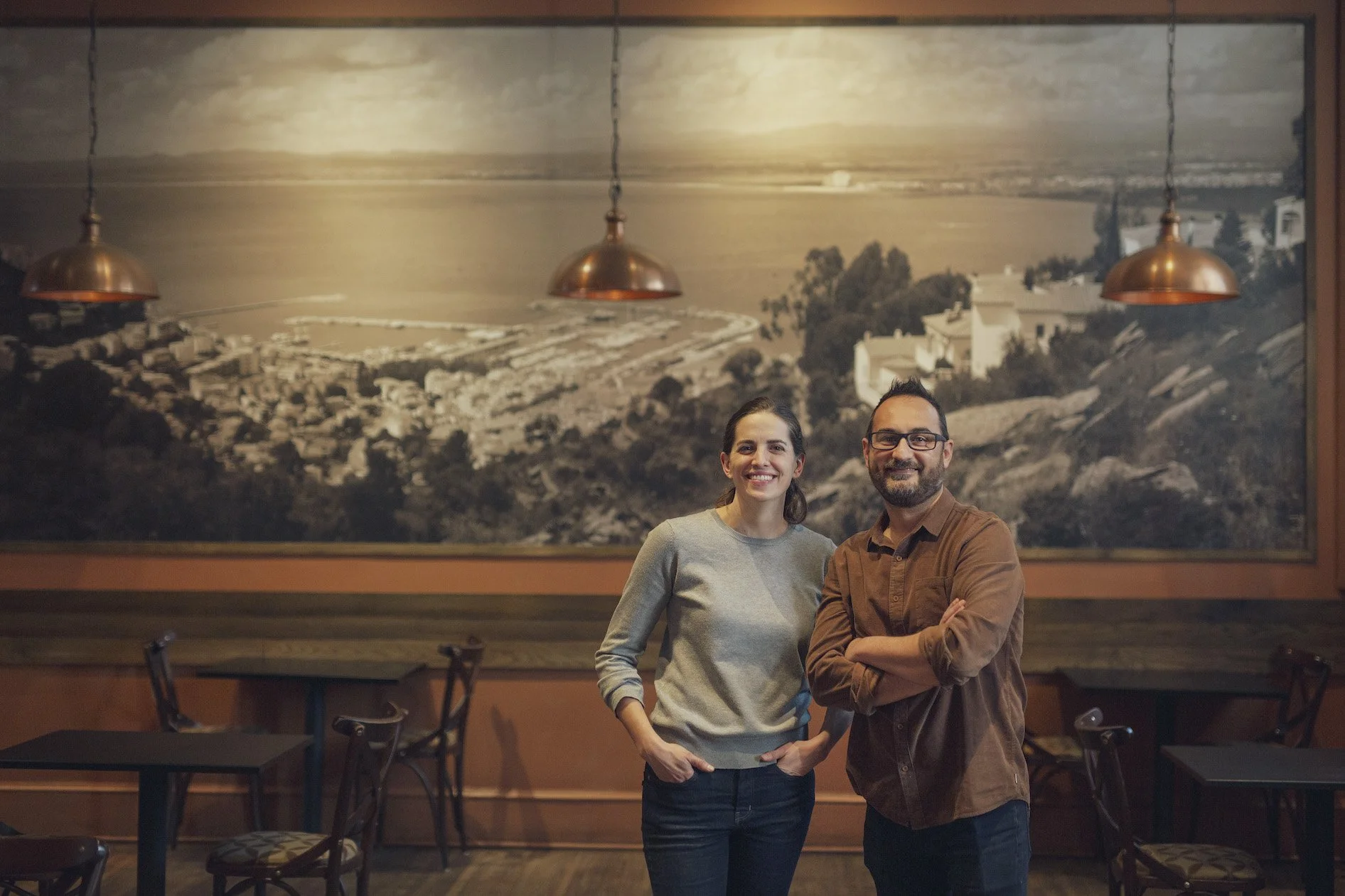 A woman and a man standing side by side in a restaurant with a large black-and-white landscape mural behind them. The woman is wearing a gray sweater and dark pants, and the man is wearing glasses and a brown shirt with rolled-up sleeves. They are smiling and posing for the photo.