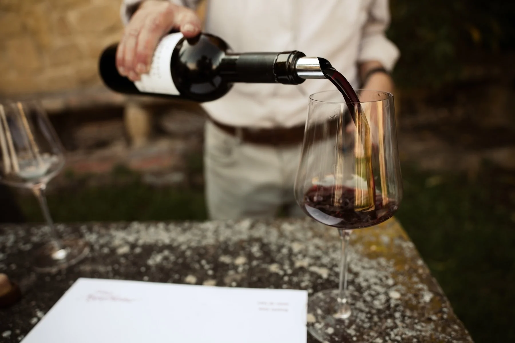 A person pouring red wine into a wine glass outdoors on a stone surface.