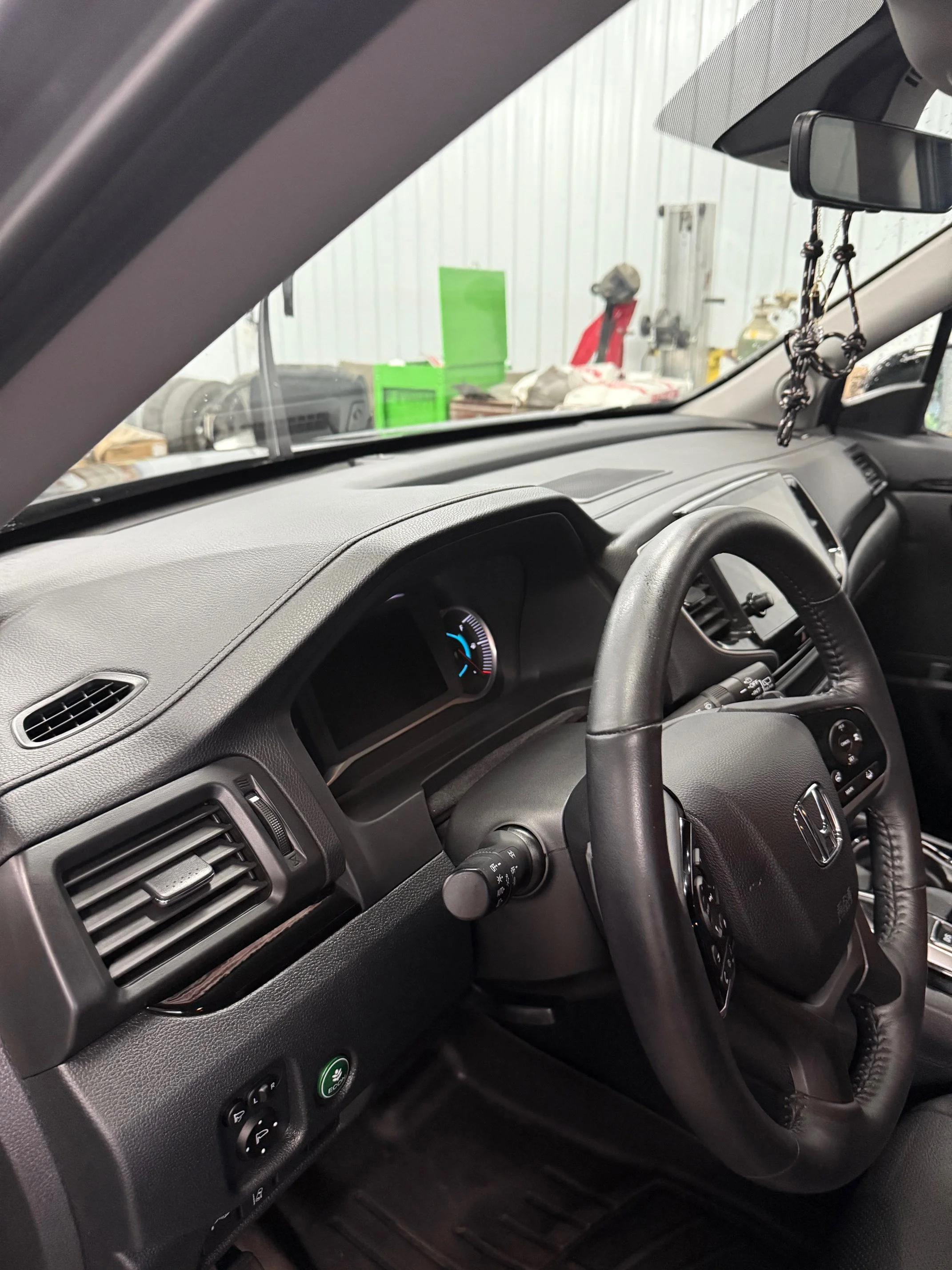 Interior of a Honda vehicle shown from the driver's side, with the steering wheel, dashboard, and part of the windshield visible. The background outside the vehicle shows a warehouse or garage with various tools and objects.