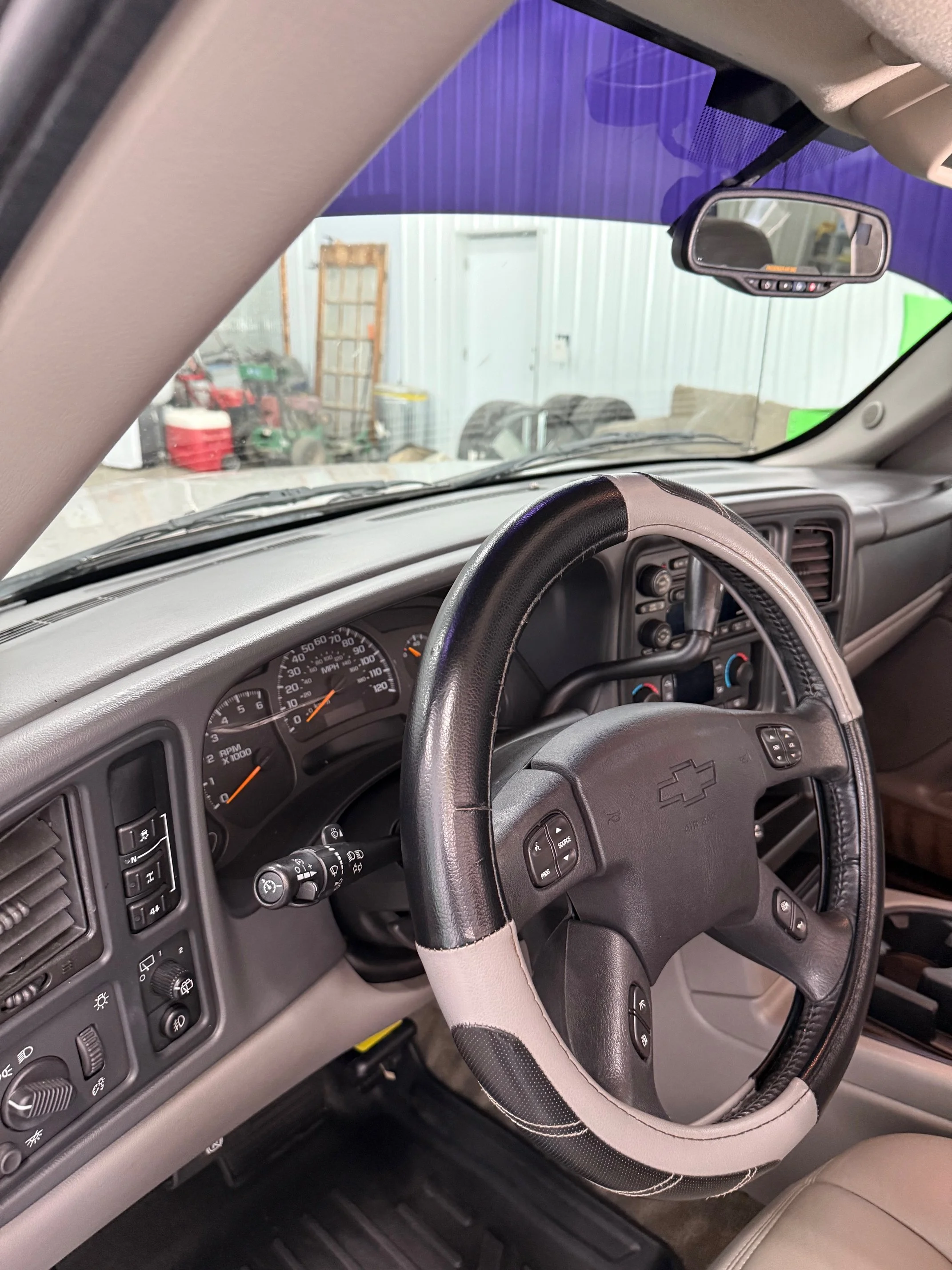 Interior of a Chevrolet truck's dashboard showing the steering wheel, speedometer, and controls, with the view through the windshield of a storage area with tools and equipment.