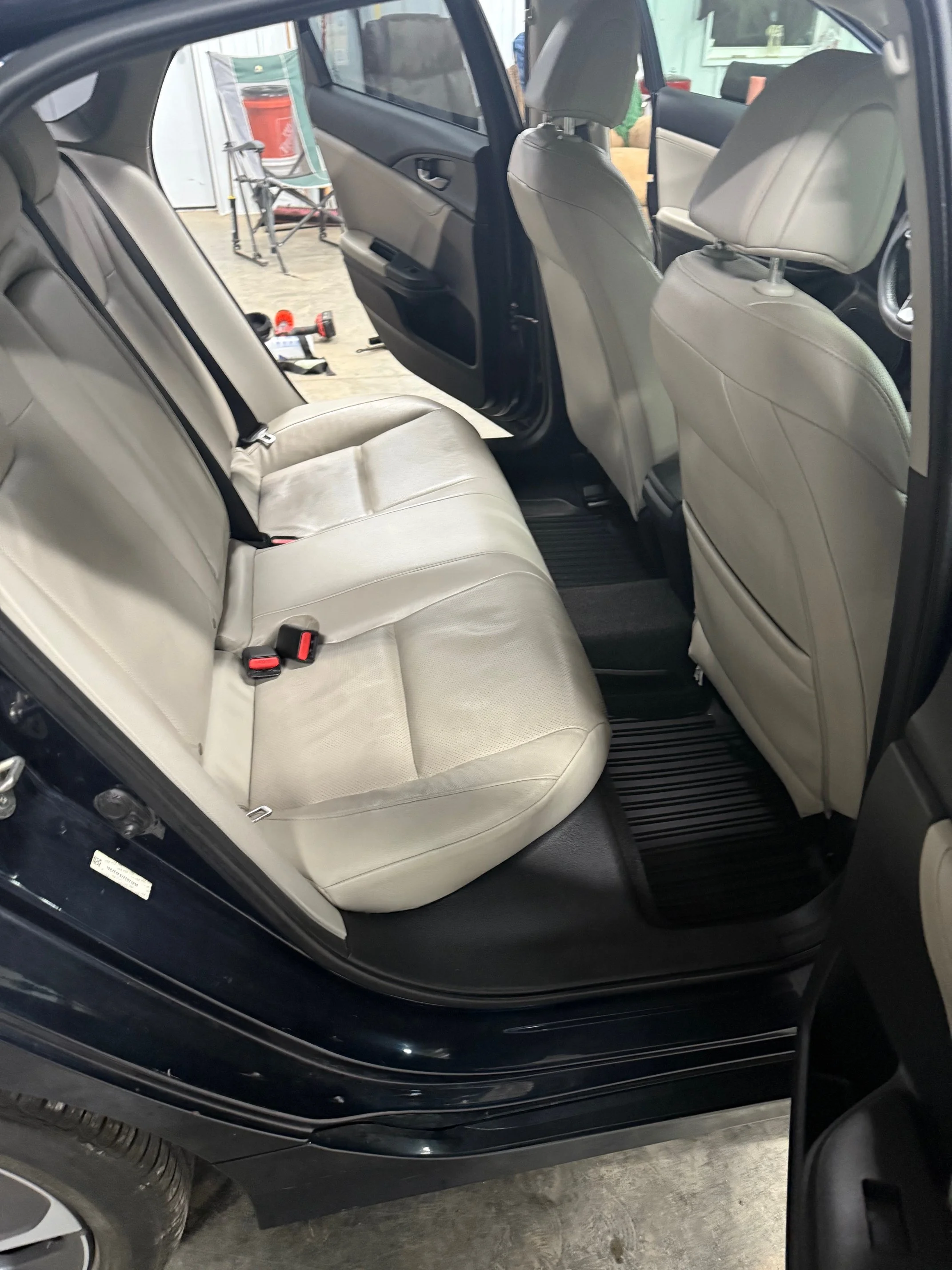 The interior of a car showing beige leather back seats, front seatbacks, and black floor mats, with an open rear door.