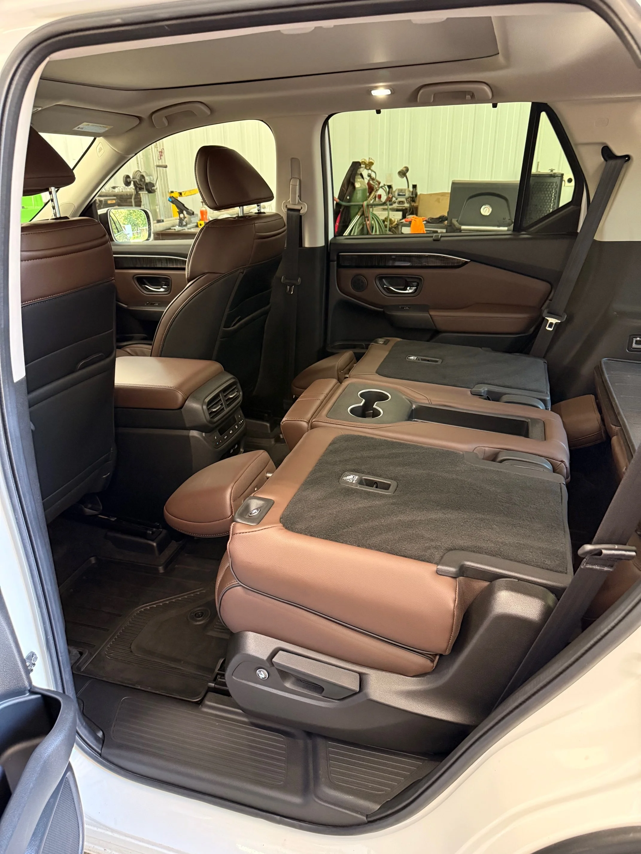 The interior of a vehicle with folded rear seats, brown leather upholstery, and the back doors open, revealing the interior and part of a garage or storage space in the background.