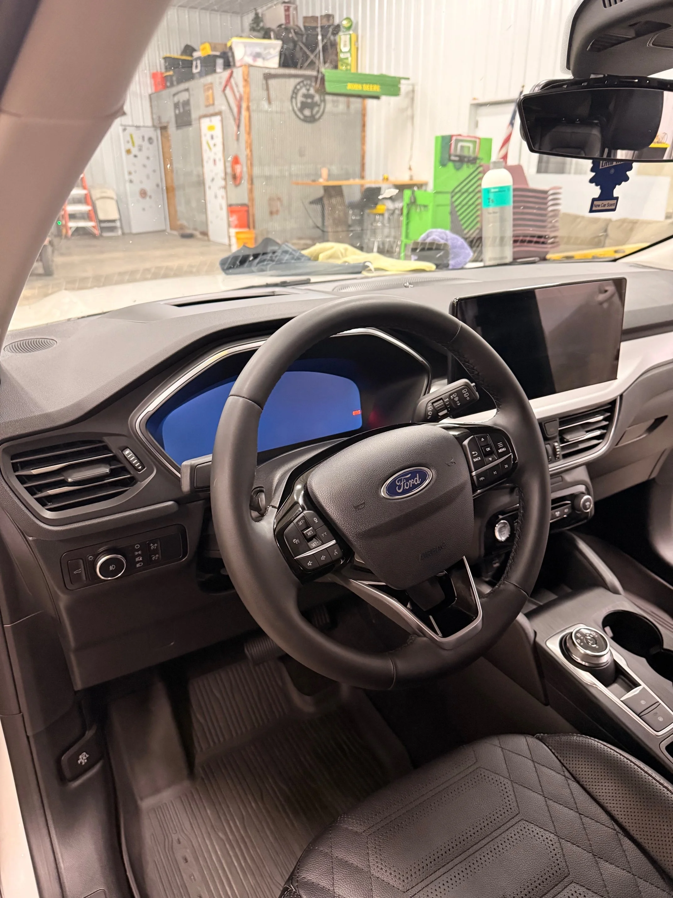 Inside view of a Ford vehicle's steering wheel and dashboard, with a large digital screen and control buttons, parked inside a garage with various tools and equipment visible outside.