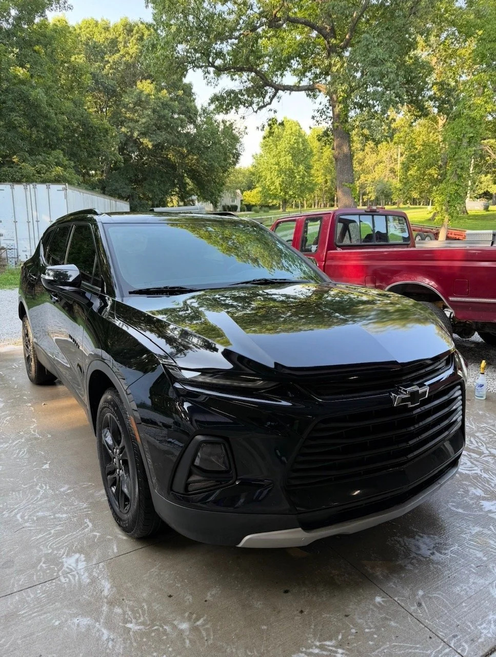 A black Chevrolet SUV parked on a driveway with a red pickup truck in the background, surrounded by trees and greenery.