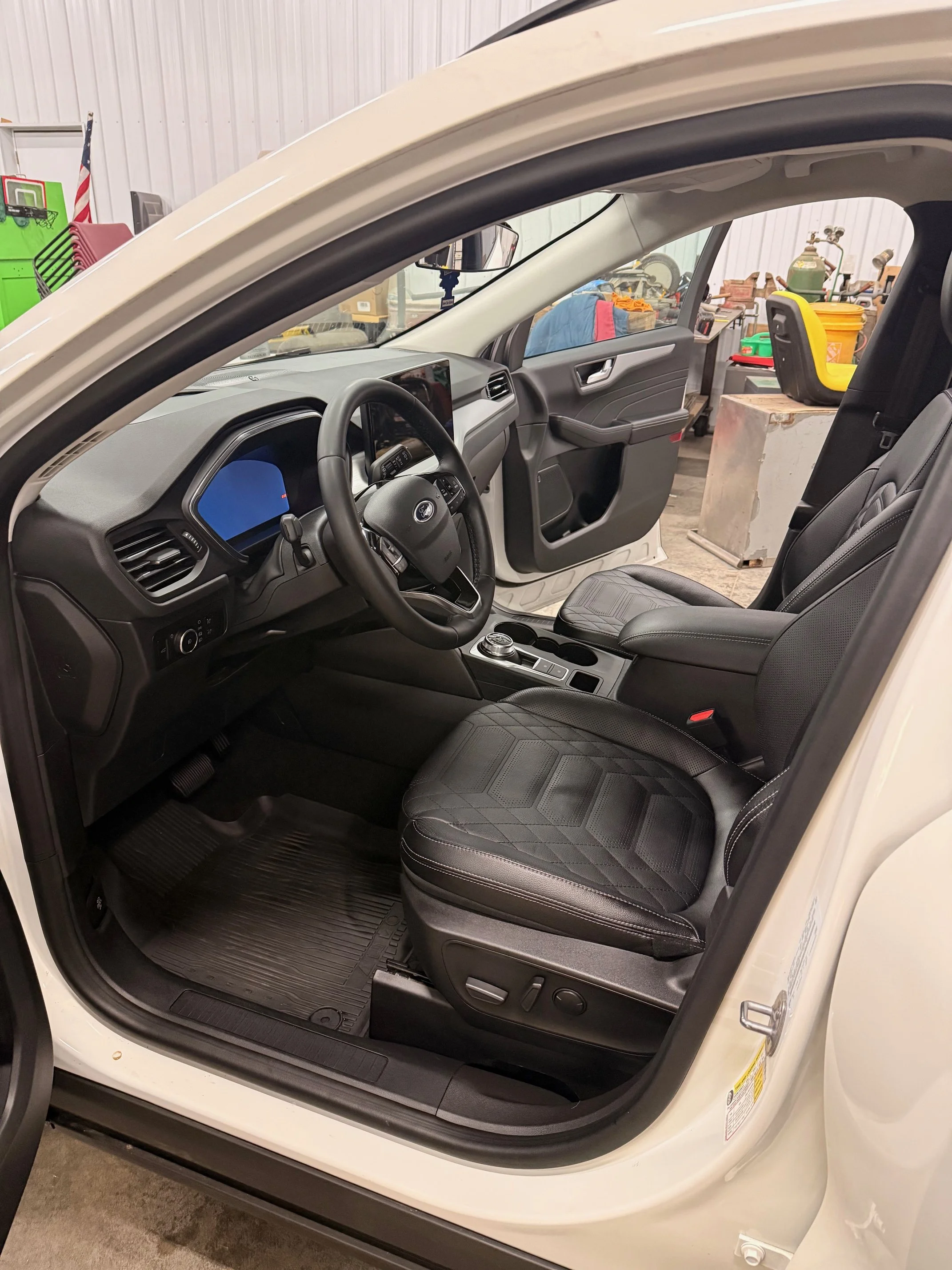 The interior of a white vehicle showing the driver's seat, steering wheel, dashboard, and front passenger seat in a workshop setting.