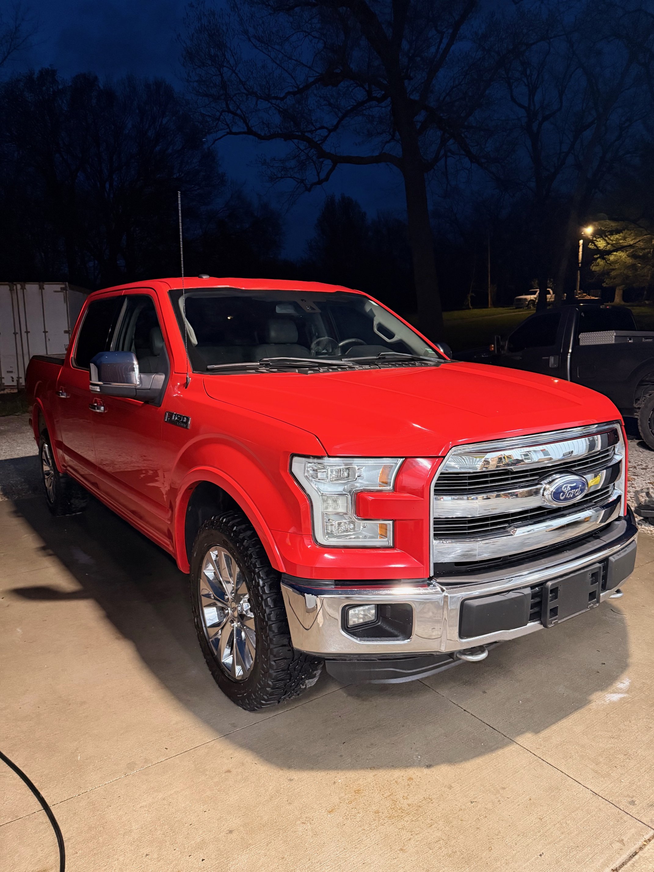 Red Ford F-150 pickup truck parked on driveway at night with trees and other vehicles in the background.
