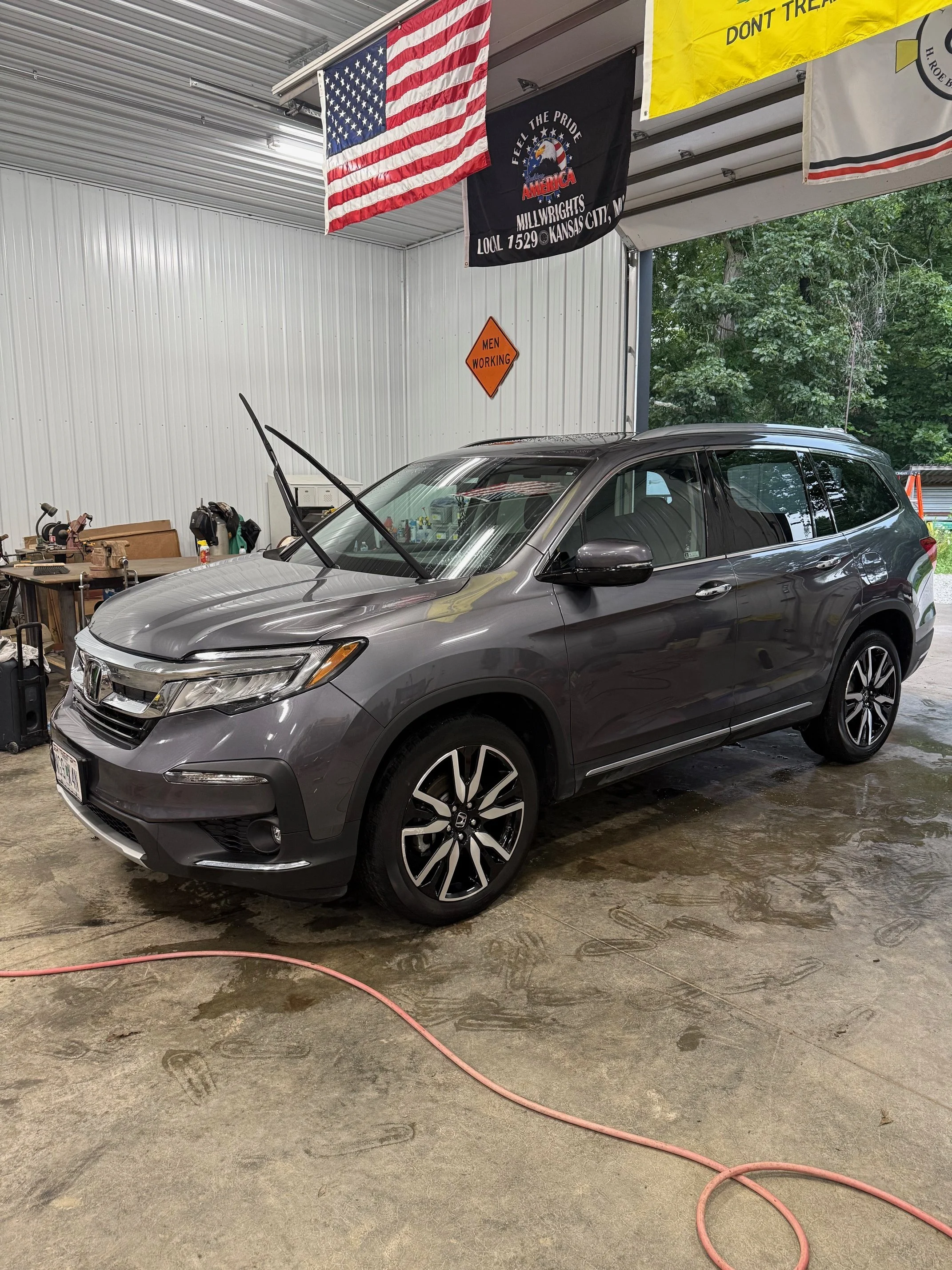 A gray Honda SUV parked inside a garage or workshop with American flags and banners hanging from the ceiling. The garage has a concrete floor with water and a pink hose on the ground. There are tools and equipment on a workbench in the background.