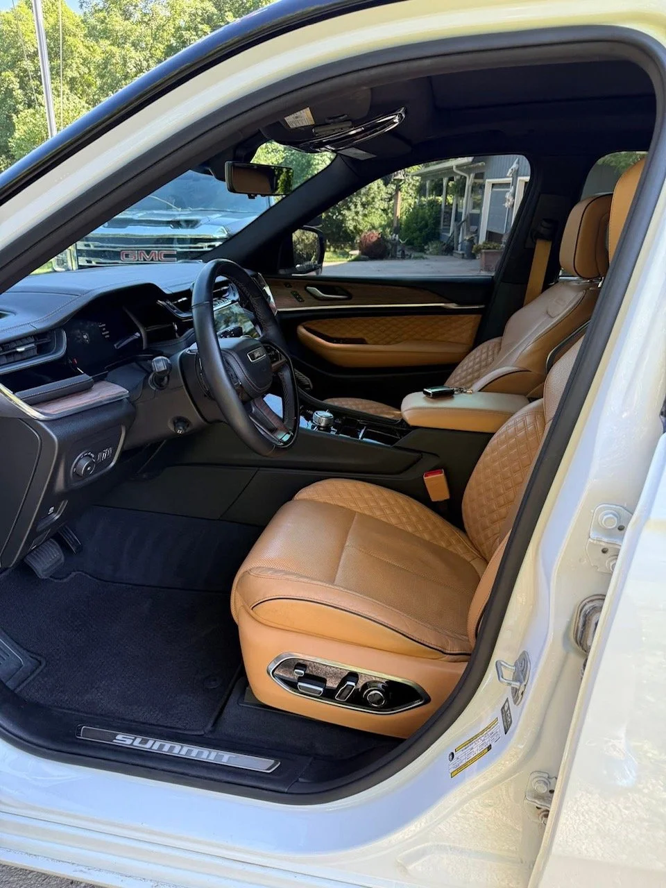Interior of a luxury vehicle with tan leather seats, black dashboard, and wood trim. The front seats are equipped with electronic controls, and there is a key fob on the passenger seat. The vehicle is parked outdoors near a house with greenery in the