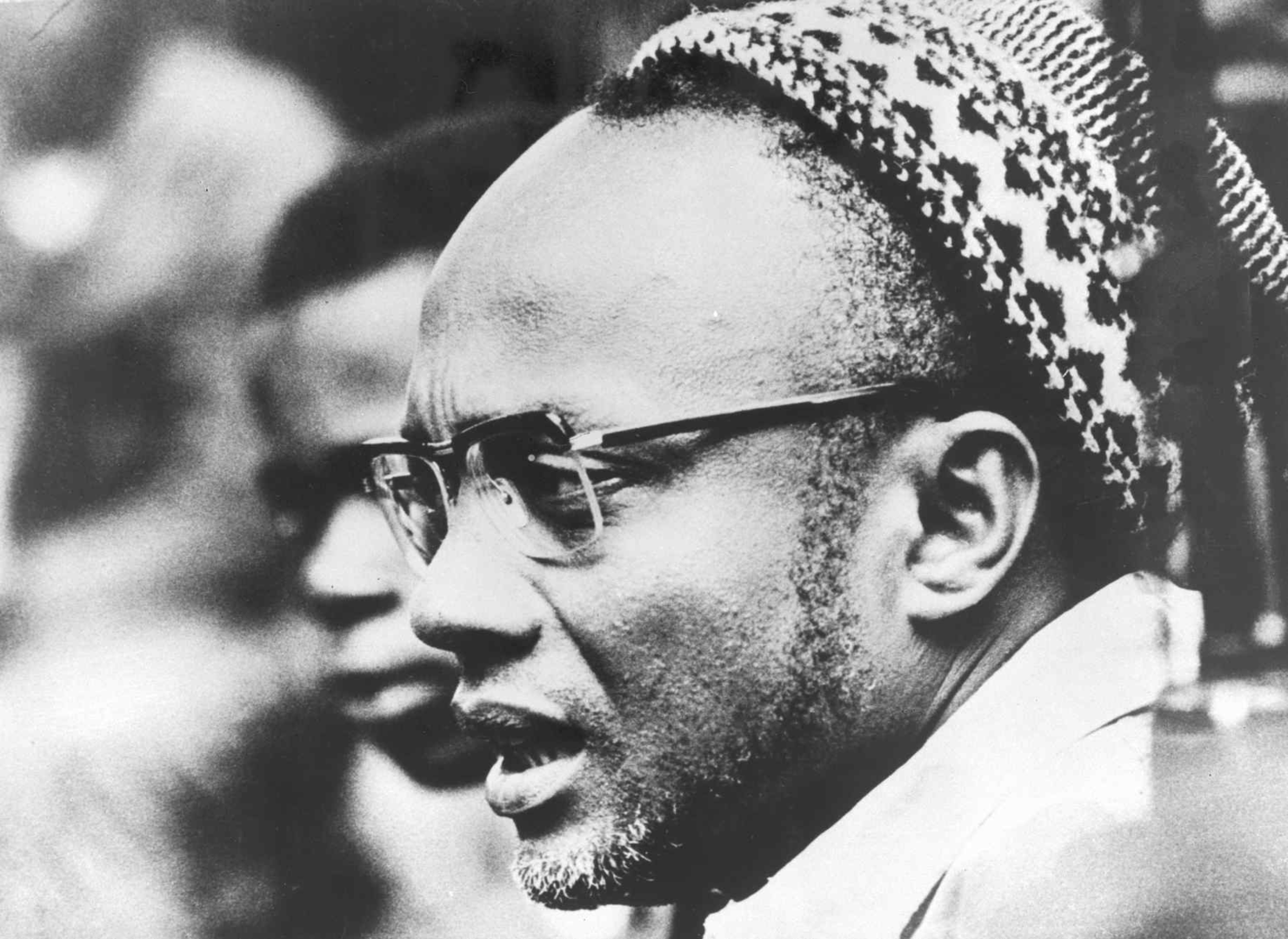 Black and white photograph of Amilcar Cabral with closely shaved hair wearing eyeglasses and a patterned headscarf, looking down and to the right.