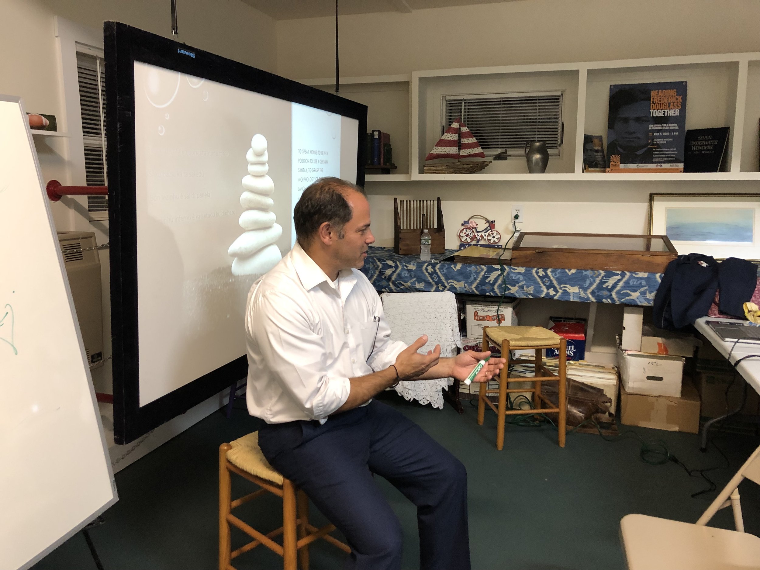 Man in white shirt sitting on a chair, gesturing with his hands, during a presentation or lecture in a room with a large screen and bookshelves.
