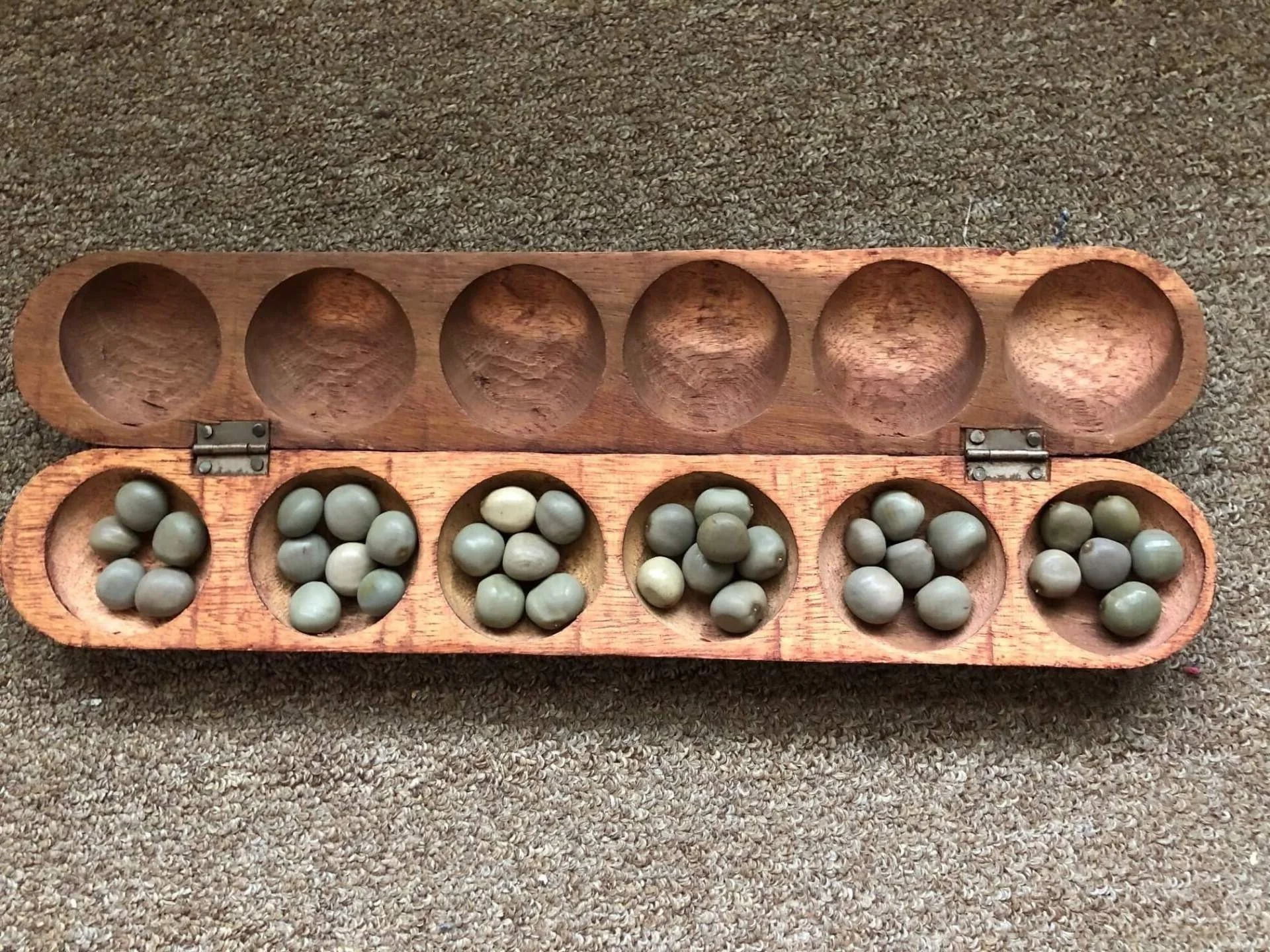 A wooden board game with six holes on the bottom filled with small, round, light-colored balls, and six empty holes at the top.