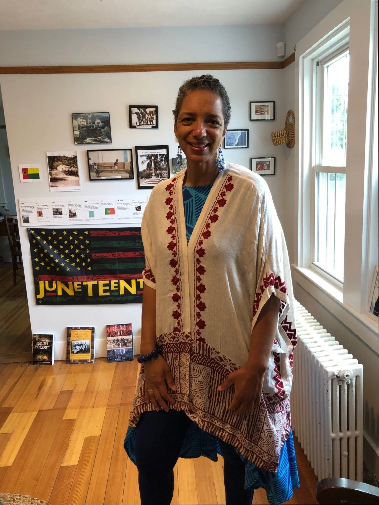 A woman standing indoors in front of a display about Juneteenth, wearing traditional African attire, with a window to her right and framed pictures on the wall behind her.