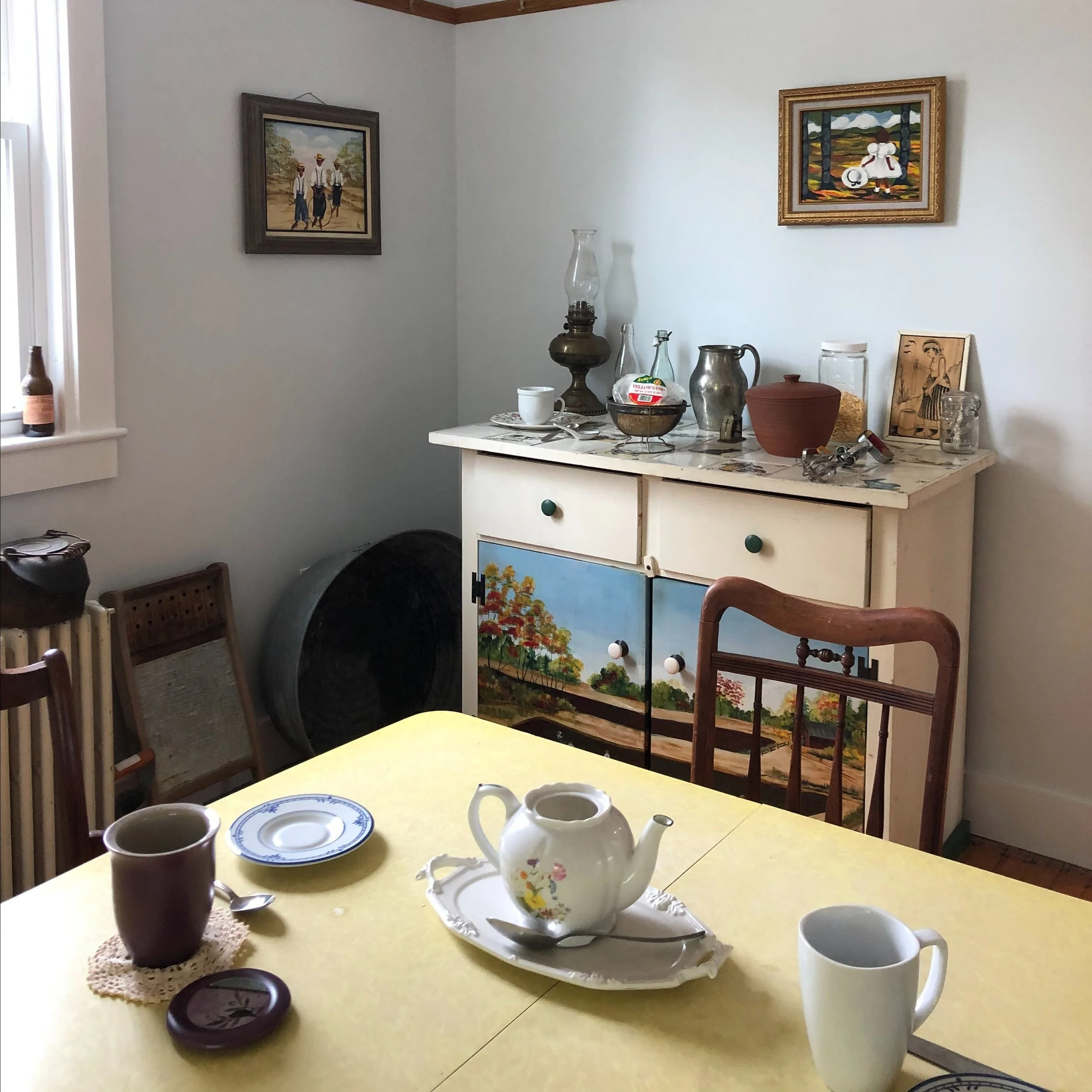 A cozy dining room with a yellow table set for tea, featuring a white teapot, a mug, a small plate, and a spoon. In the background, there is a white sideboard decorated with vintage items and framed artwork on the wall.