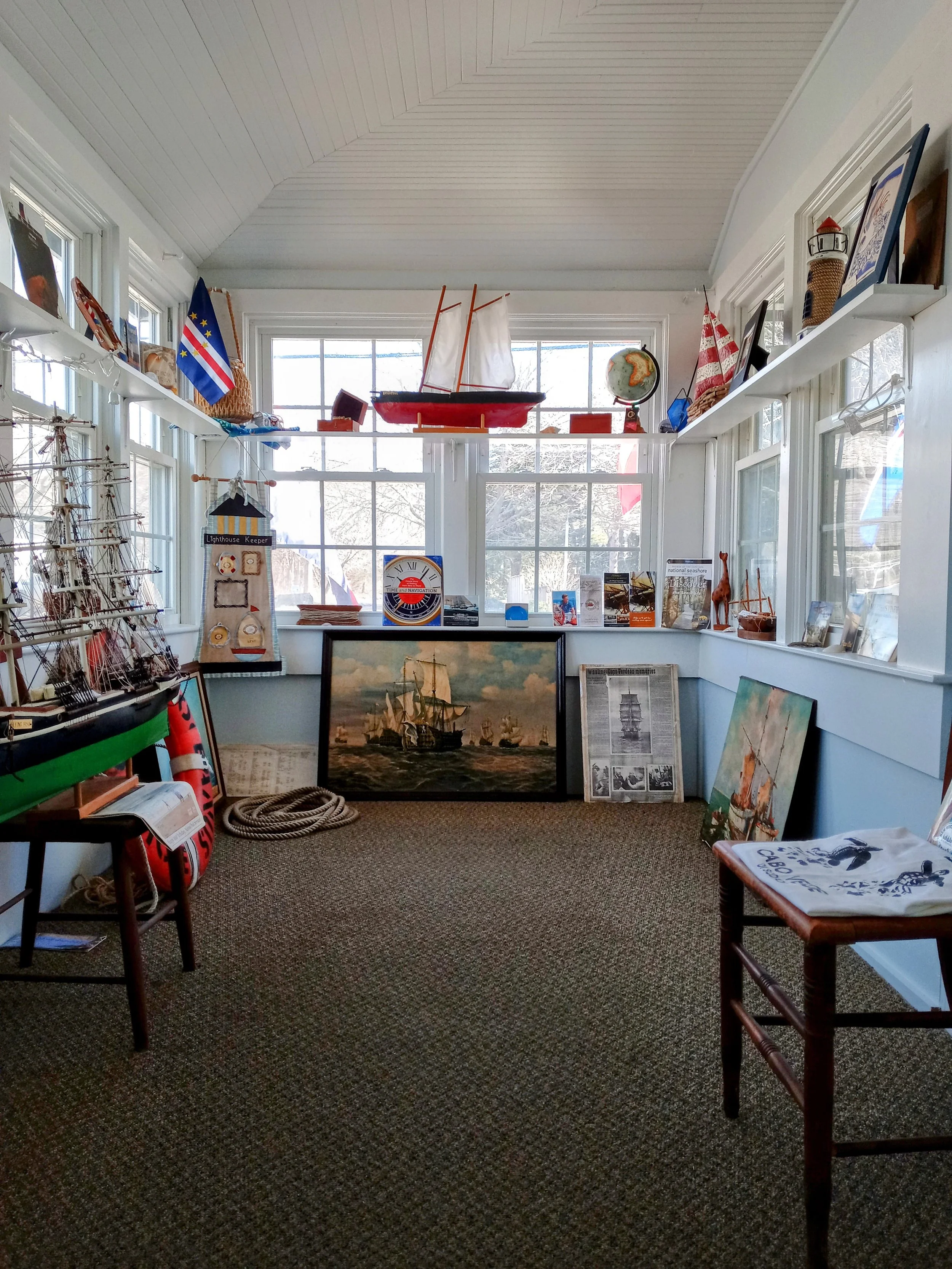 A nautical-themed room with model ships, marine paintings, flags, a globe, and maritime decor displayed on shelves and the floor.