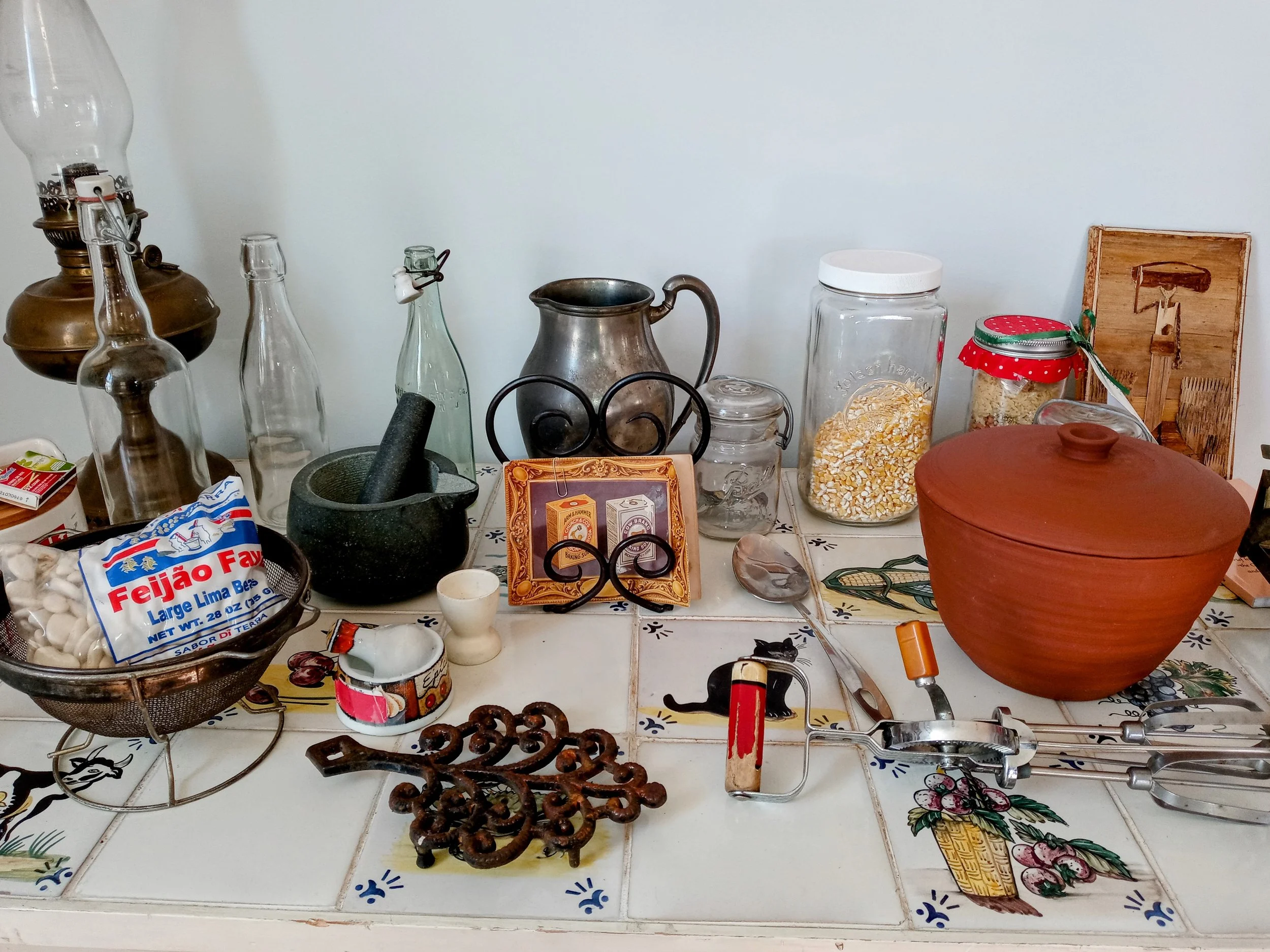 A cluttered kitchen countertop with various glass bottles, jars, a mortar and pestle, a large brown ceramic container, a spoon, a small black cat figurine, and miscellaneous kitchen items and decorations.