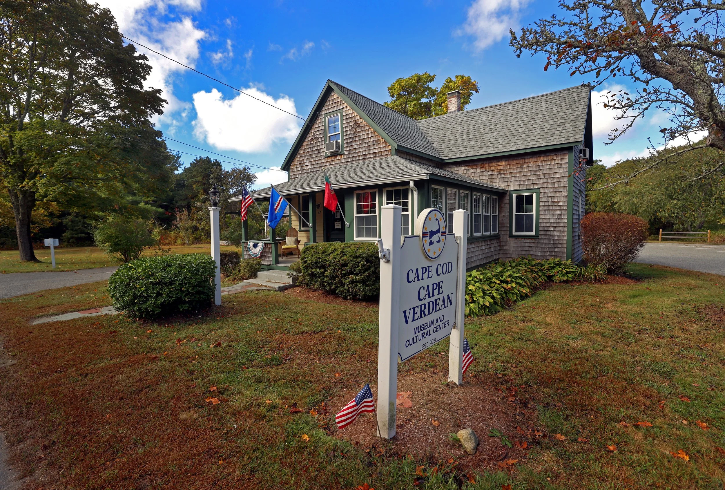 A historic house with a gabled roof, shingle siding, and a small porch decorated with flags, located at the Cape Cod Cape Verdean Museum and Cultural Center. The yard has grass, bushes, and a sign with flags in front.